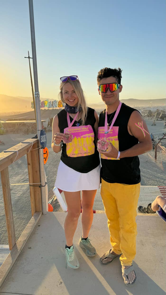 A man and a woman, both wearing black shirts with neon graphics, pink medals, and sunglasses, standing on the top of the BRC ski, snowboard, and sledding slope at Burning Man.