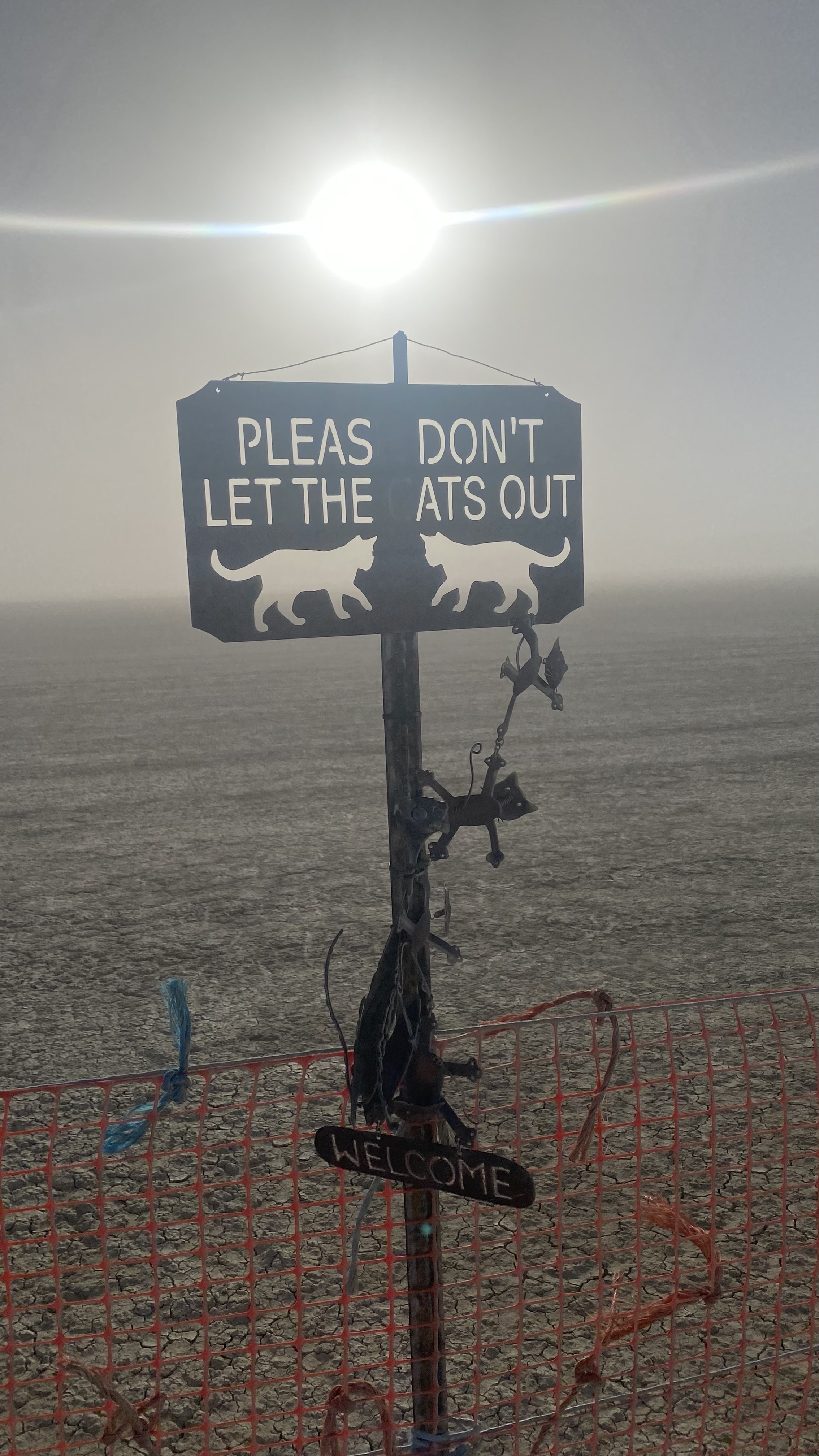 Sign on a post in a barren landscape at Burning Man reading, "Please Don't Let the Cats Out." Decorations hang from the post, including a small "Welcome" sign