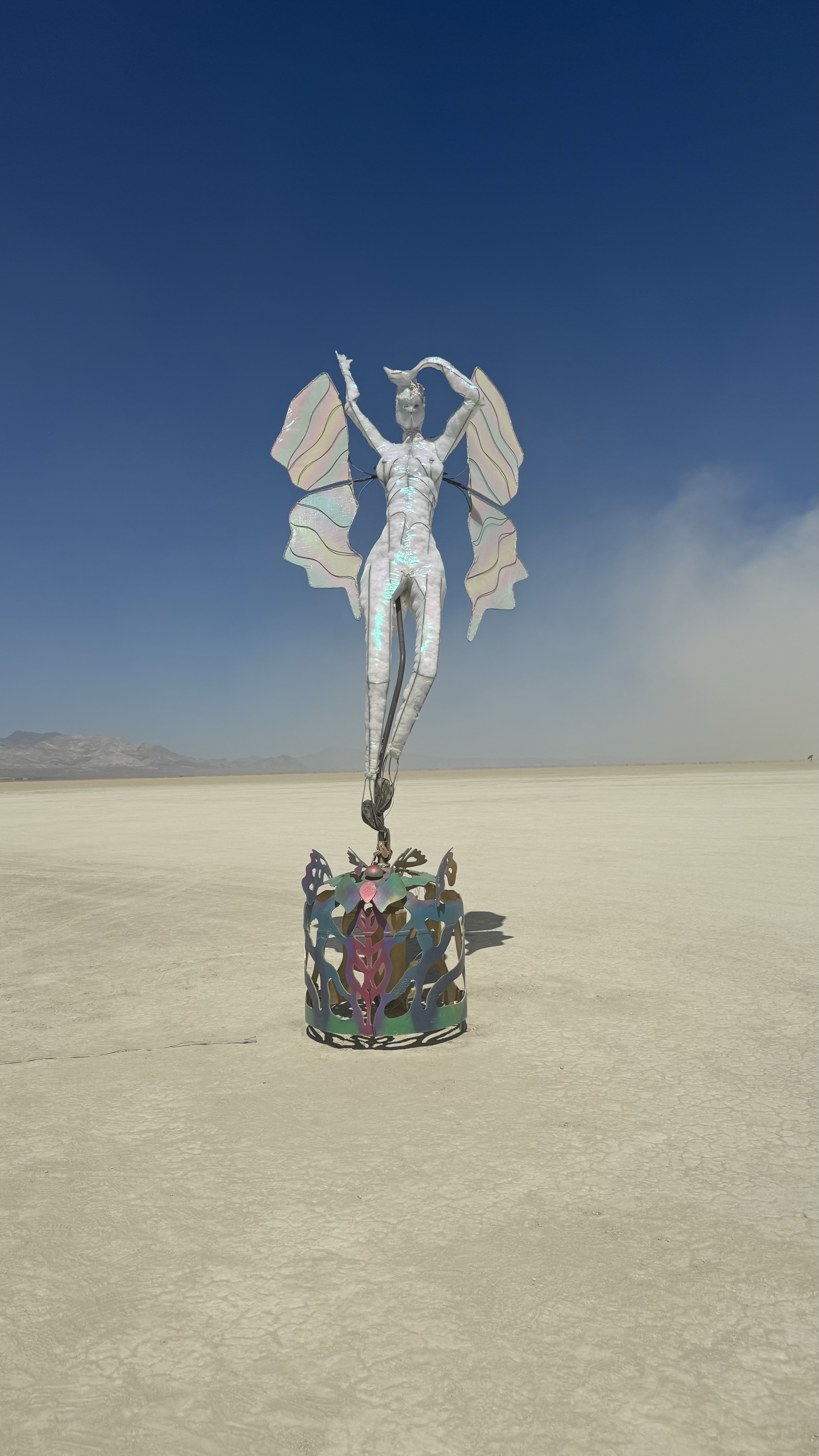 A metallic statue of a winged figure standing in a desert setting at Burning Man, with mountains visible in the distance