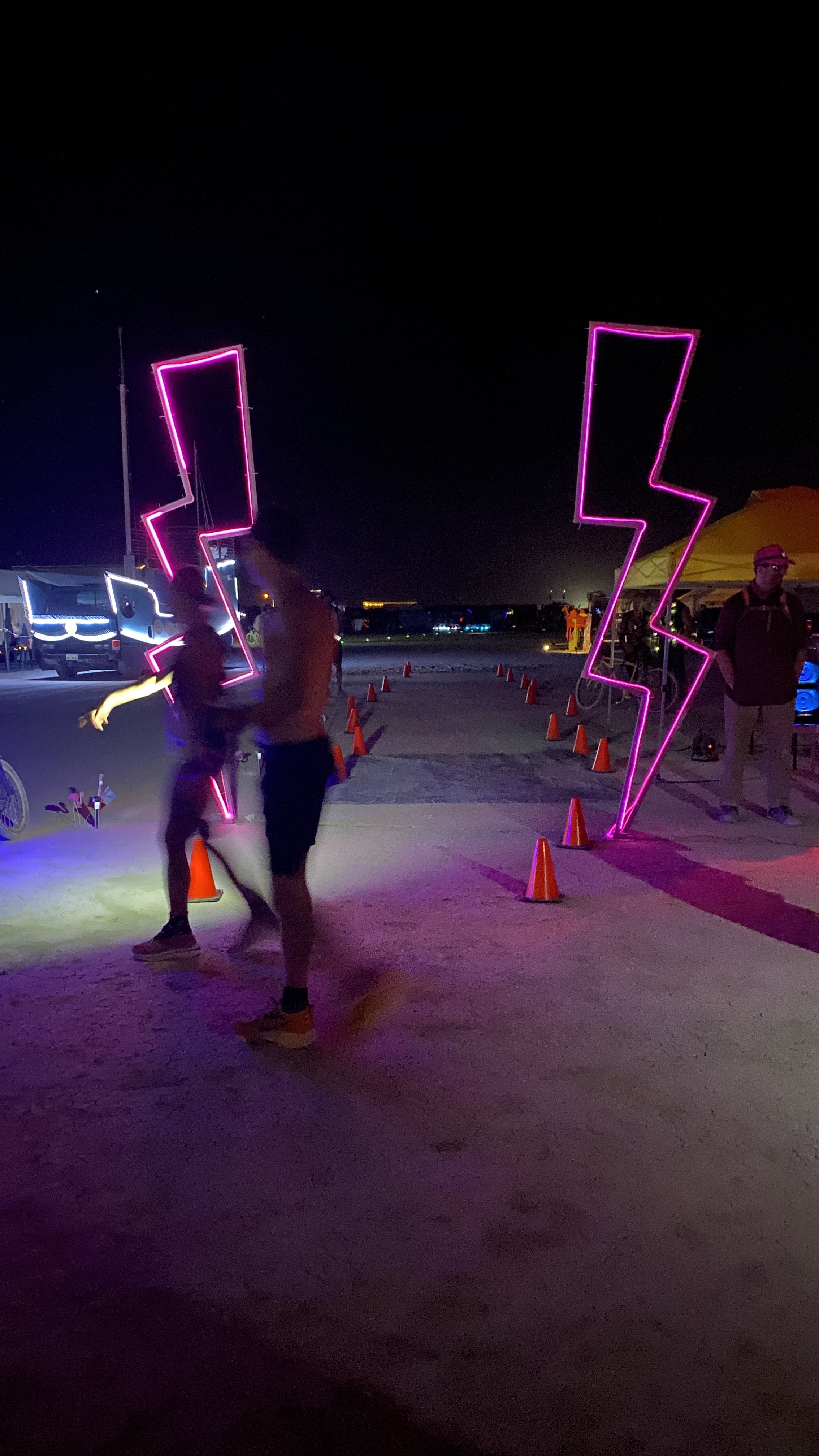 Two people walk through a lightning bolt-shaped neon installation at night with orange cones and Burning Man attendees in the background