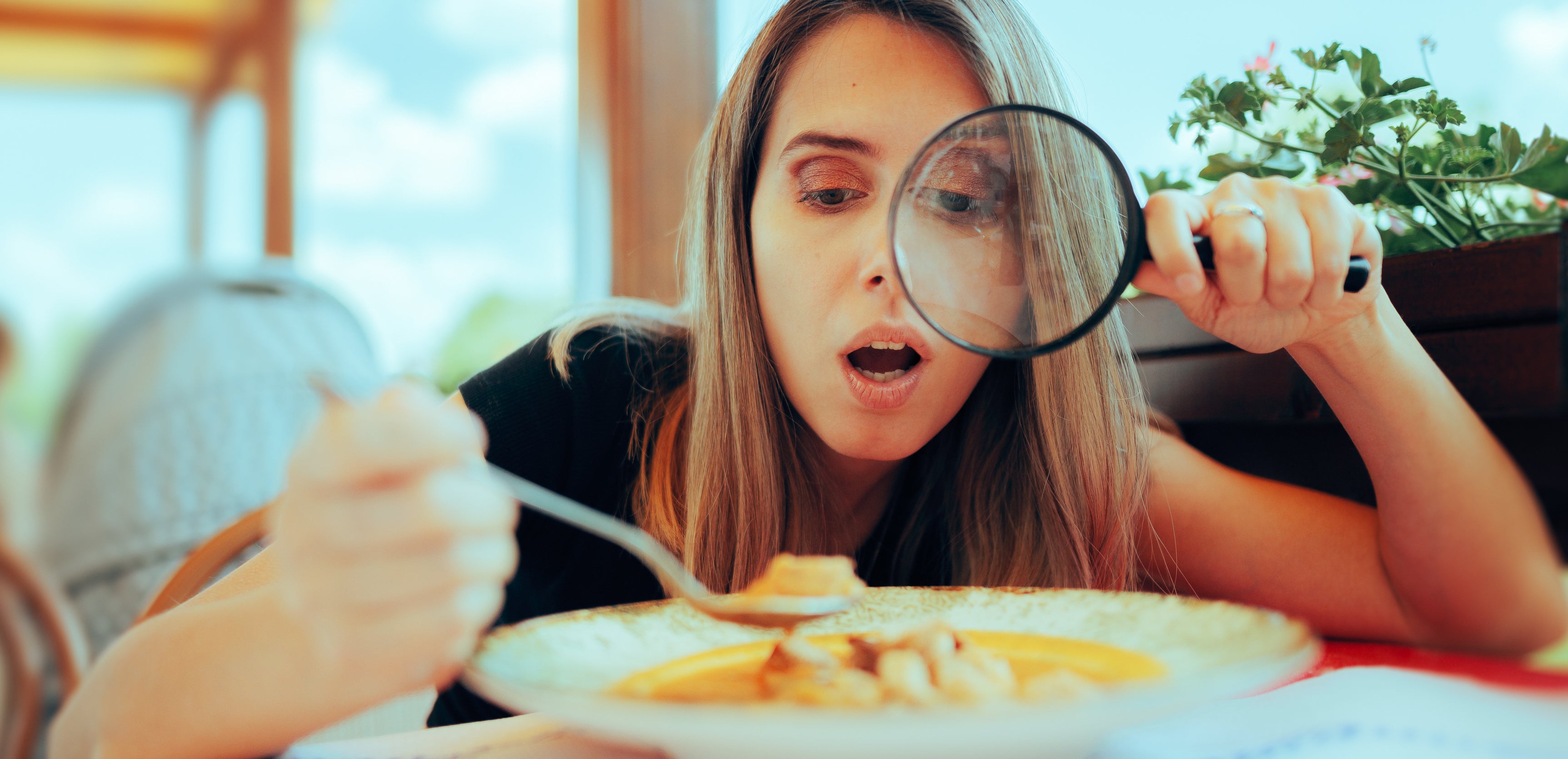 A woman in a restaurant looks surprised while examining her meal with a magnifying glass, holding a spoon in her other hand