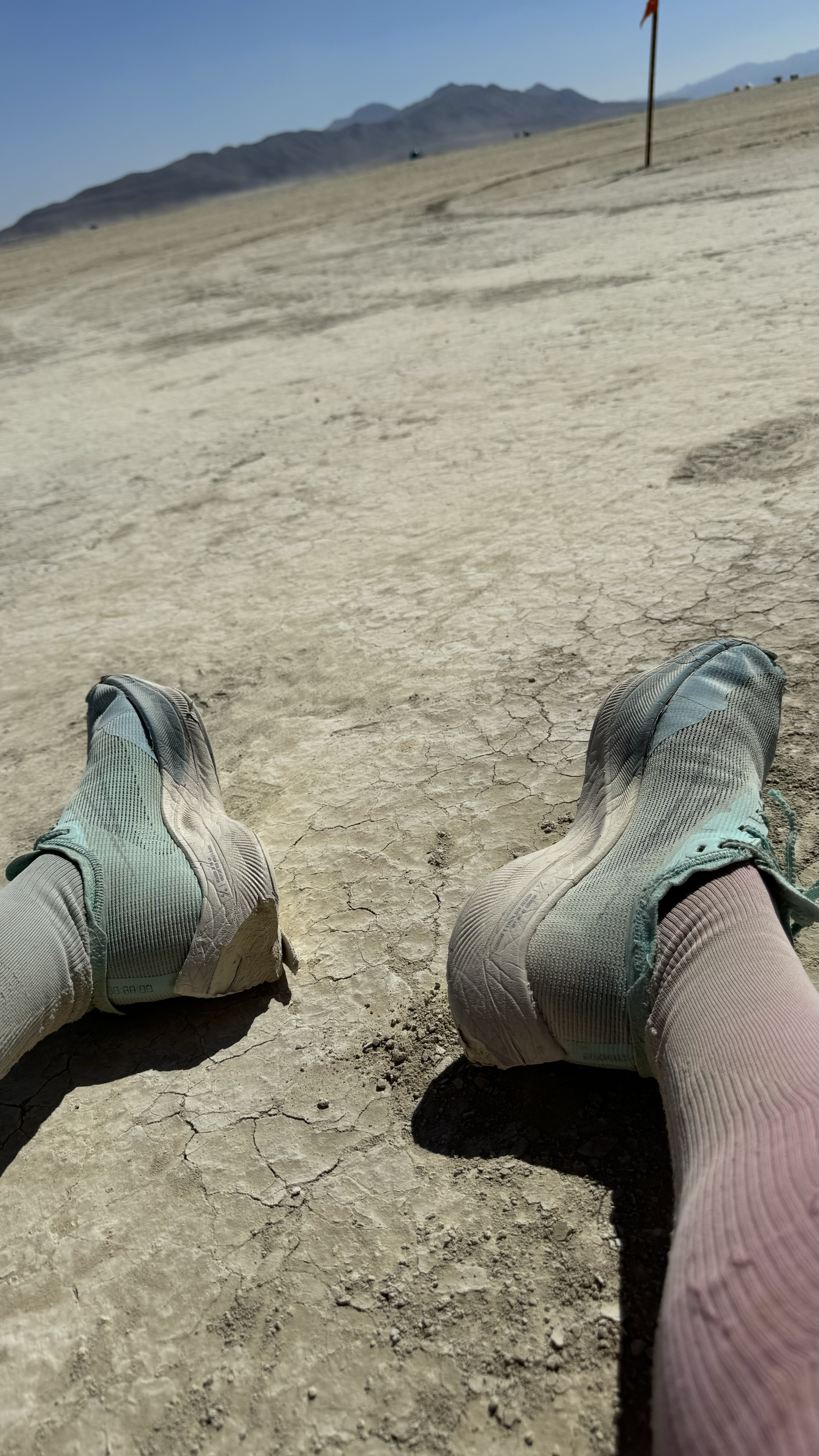 Elizabeth wearing Nike running shoes and CEP Compression socks sitting on dry, cracked ground in a desert-like area, looking out toward mountains in the distance and a small flag on the right