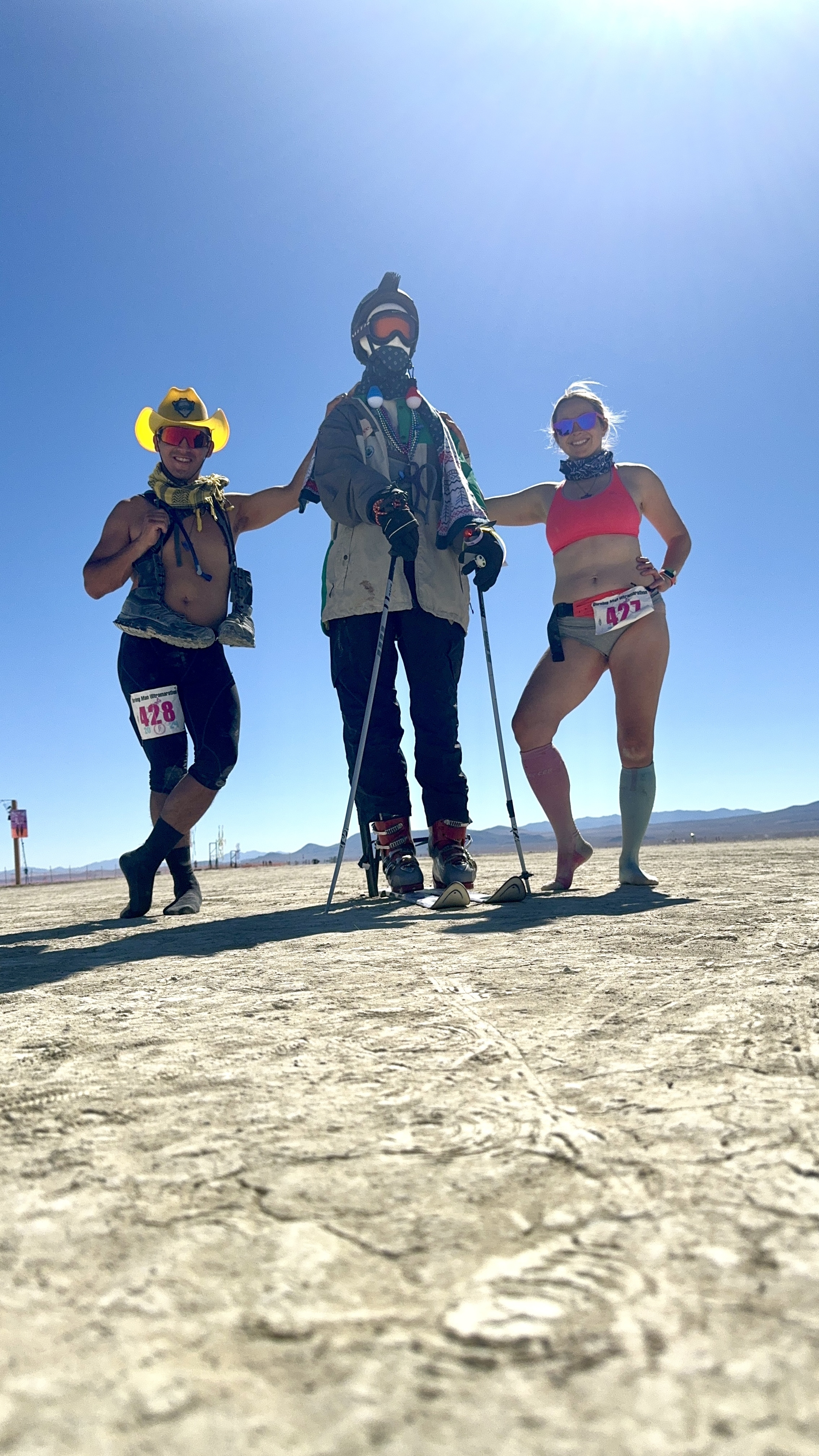 Two runners and one art figure with hiking gear using poles, pose on a flat, open landscape with race bibs.