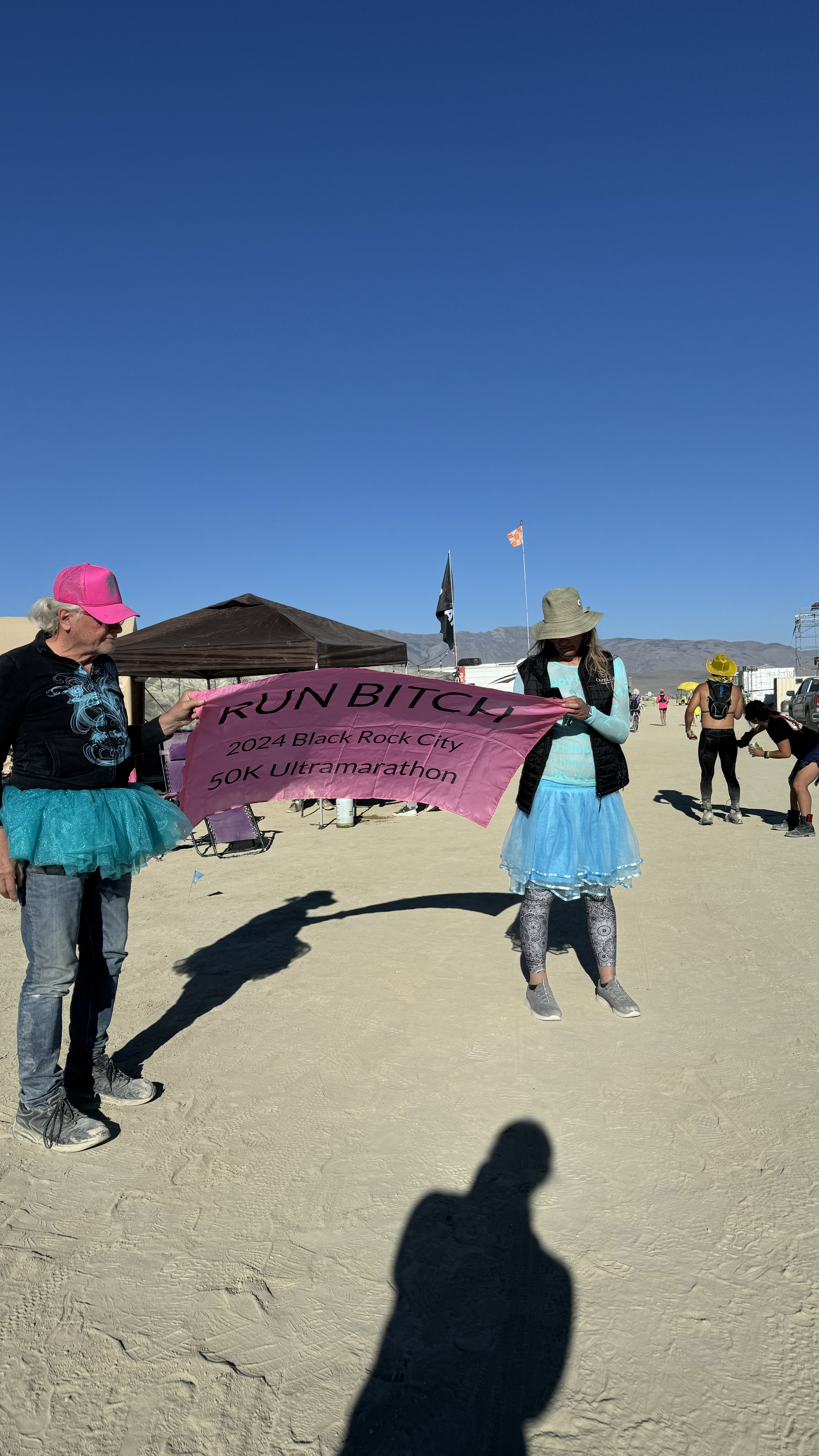 Two people in tutus hold a pink banner that reads "RUN BITCH 2024 Black Rock City 50K Ultramarathon" at an outdoor event with others in the background.