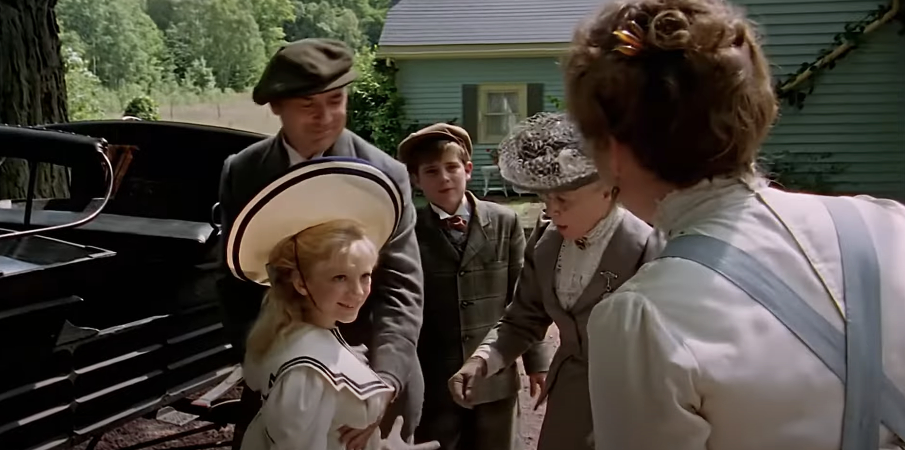 Actors in period costumes from the movie &quot;Anne of Green Gables&quot; are gathered near a vintage car in a rural setting