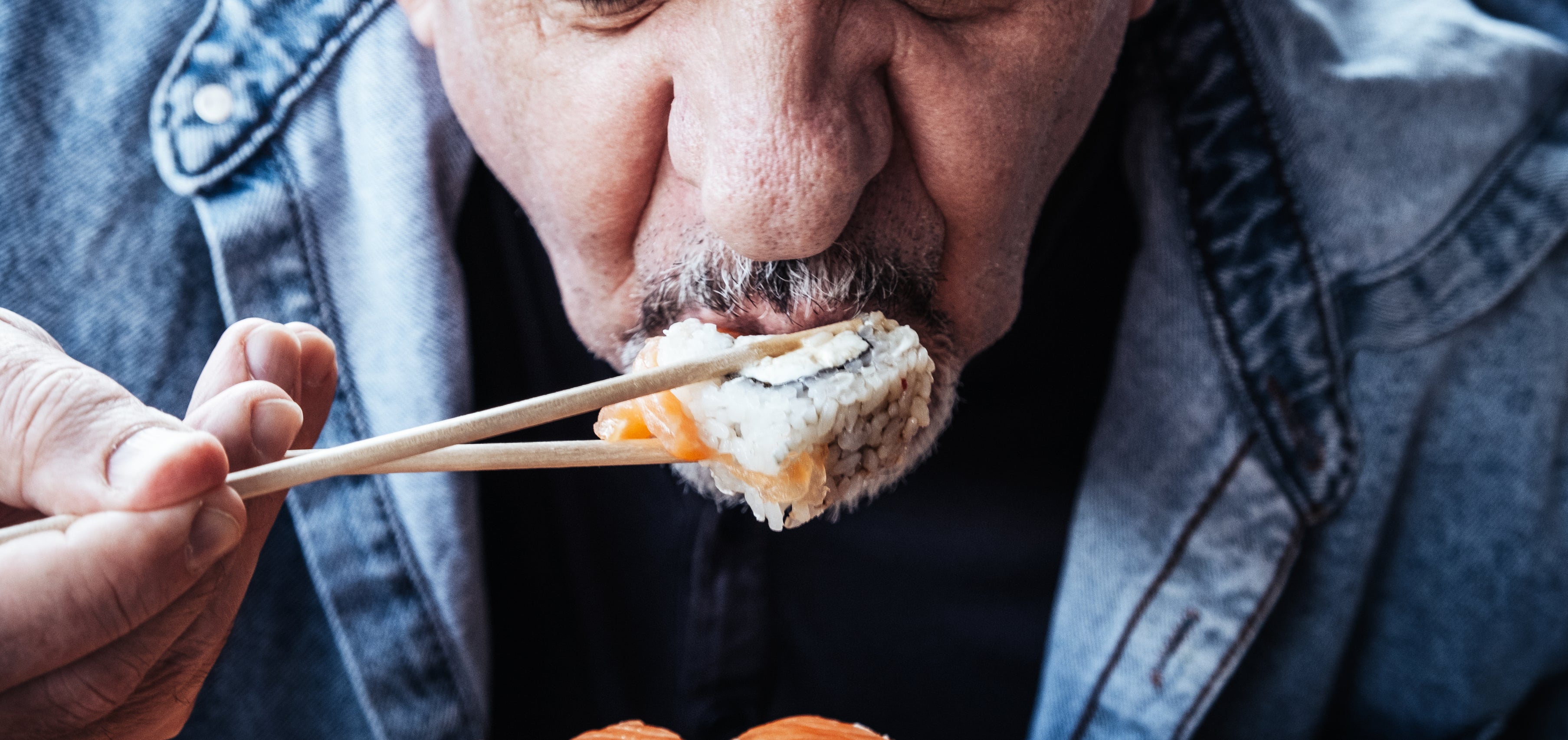 Man in denim jacket eating sushi with chopsticks
