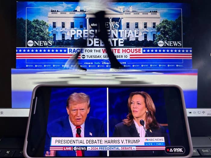 Donald Trump and Kamala Harris debate during the 2024 presidential debate, displayed on a phone screen in front of a background showing the White House and a news banner