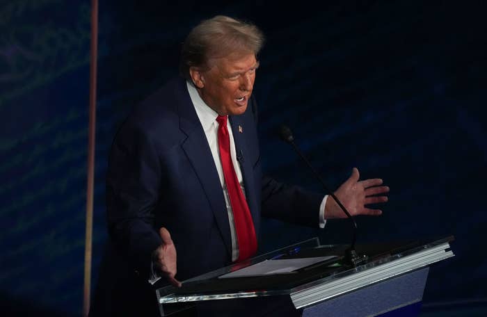 Donald Trump at a podium, speaking with gestures, wearing a dark suit, white shirt, and red tie