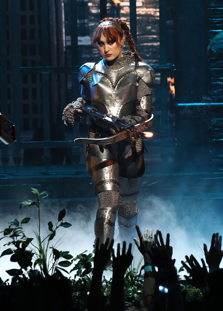 Chappell in a full metallic, medieval-style armor suit performing on stage and holding a bow and arrow, surrounded by raised audience hands and stage props resembling plants and fog