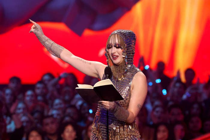 Chappell is onstage at a public event, wearing a sparkly, hooded outfit and reading from a book while pointing upwards