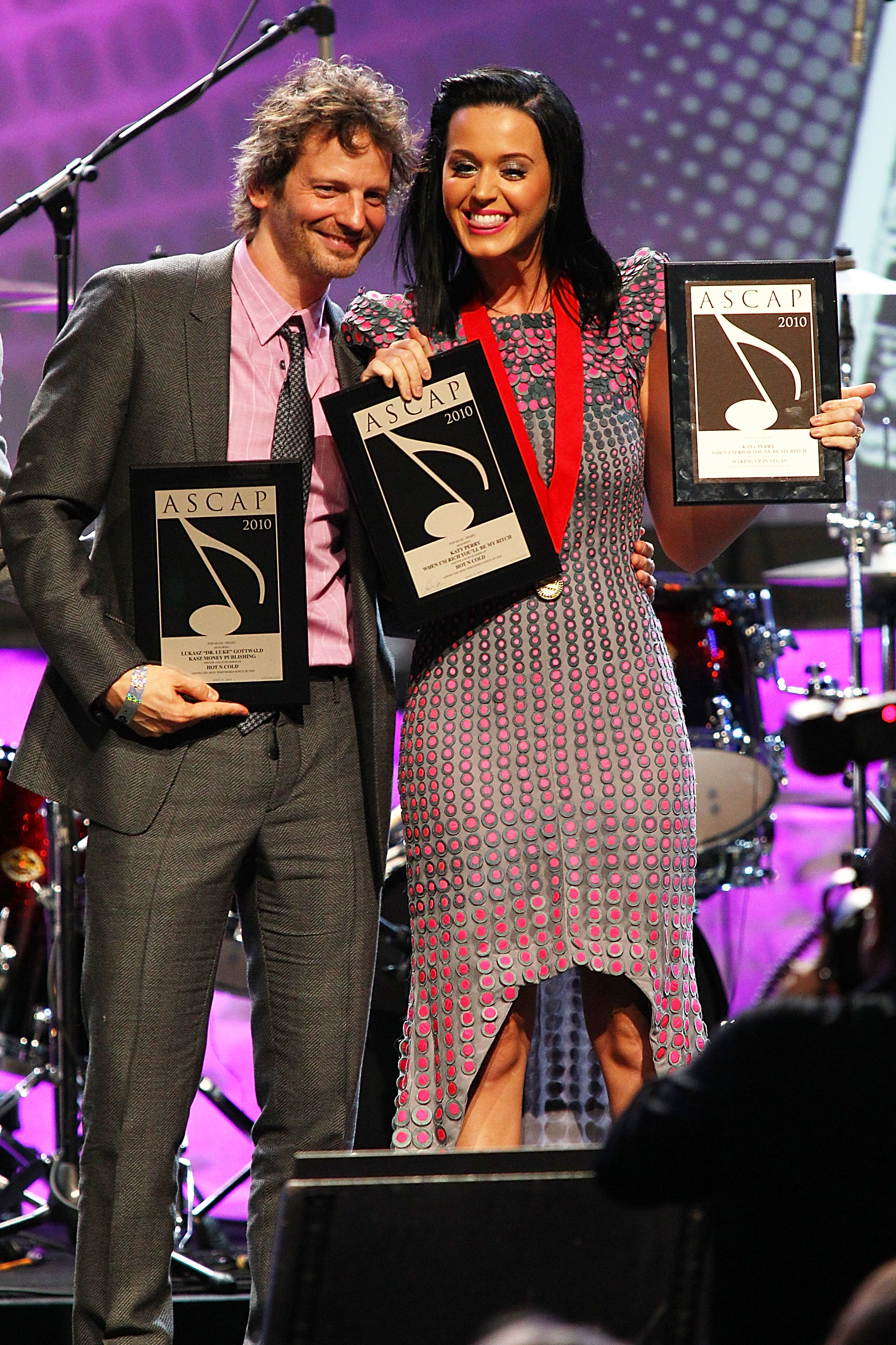 Dr. Luke and Katy Perry smiling, holding ASCAP awards on stage; Katy Perry wears a fitted patterned dress and a medal