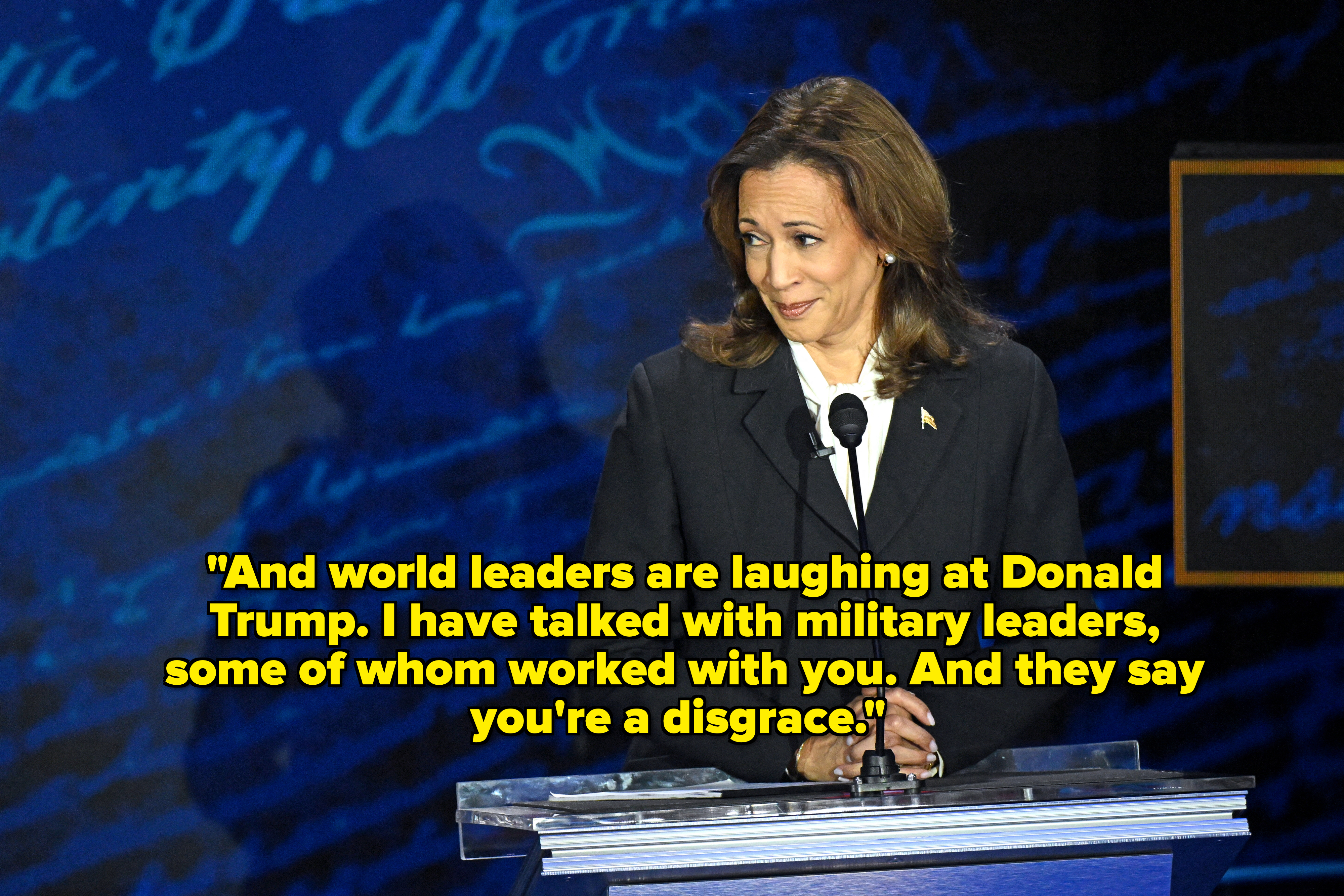 Kamala Harris stands at a podium wearing a dark suit, looking to her left with a slight smile against a blue backdrop with cursive writing