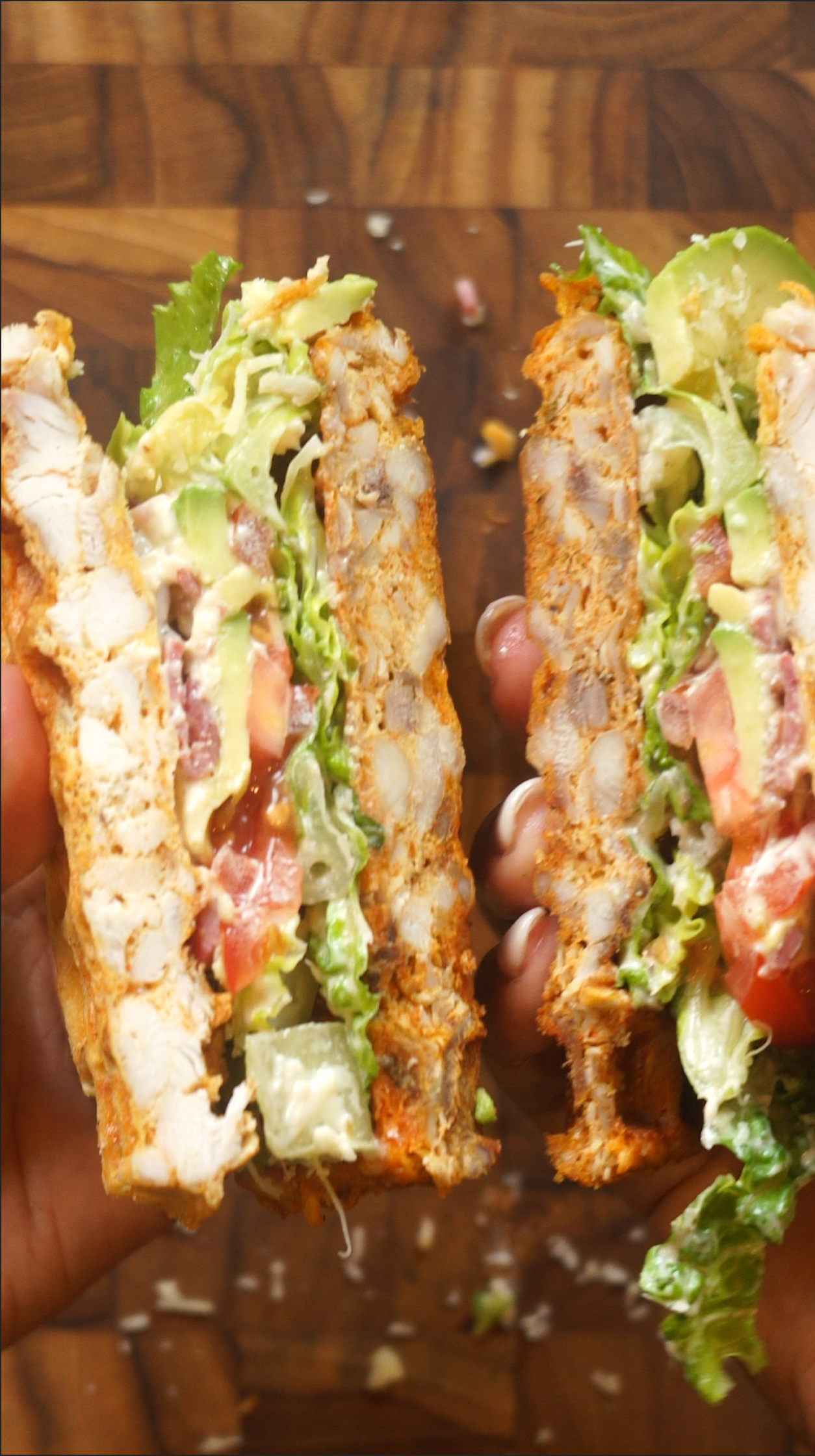 A hand holding a cut chicken sandwich filled with lettuce, tomato, avocado, and cheese. The sandwich appears to be on toasted bread
