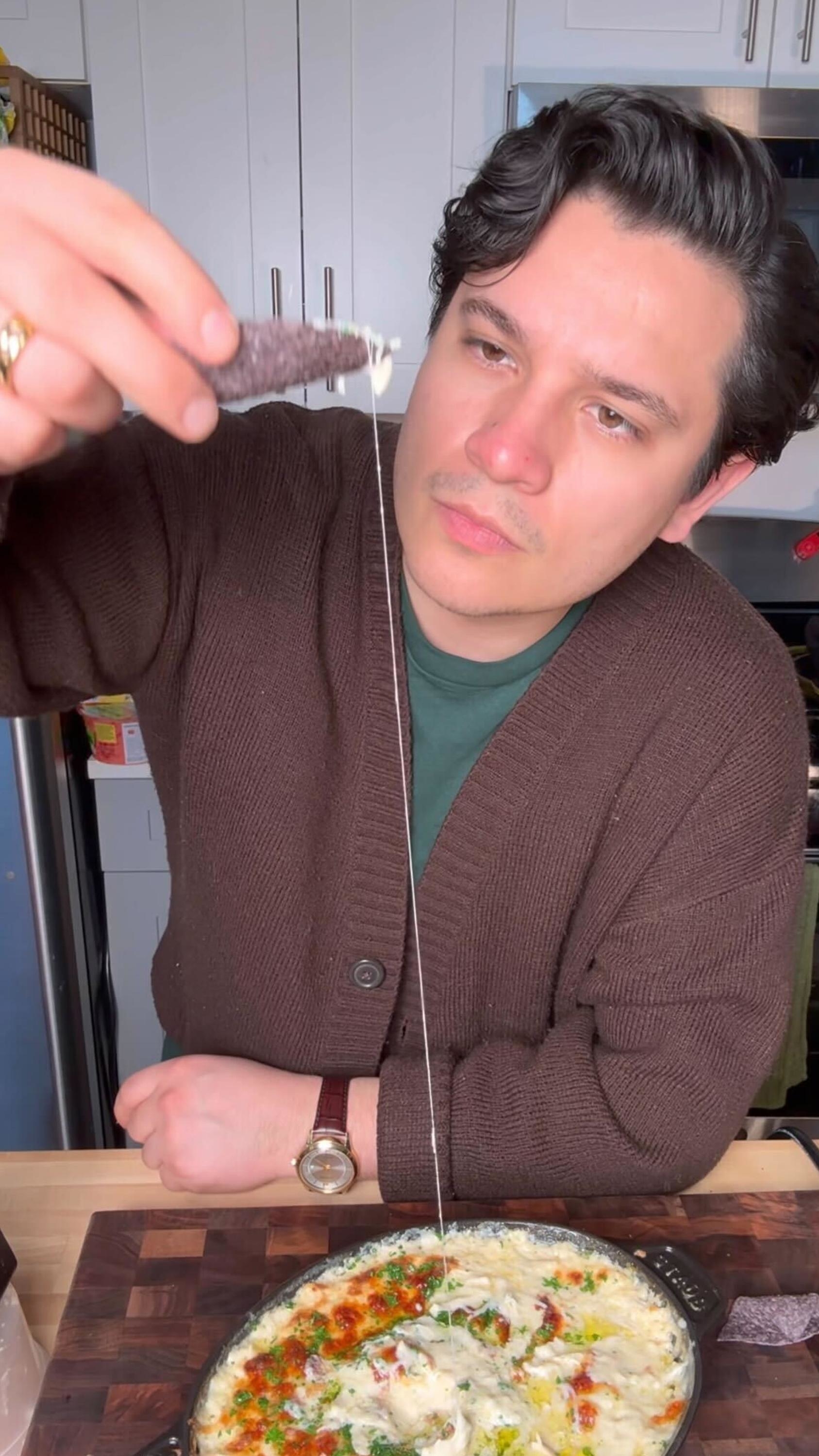 A man in a brown cardigan holds a chip above a bowl of cheesy dip, with a string of melted cheese connecting them