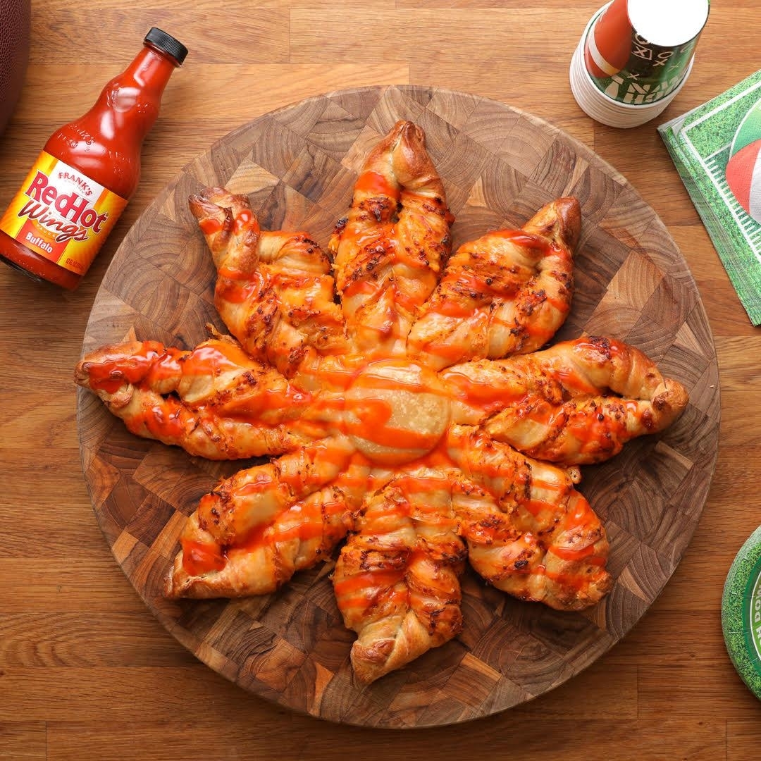 A star-shaped pizza bread covered in cheese and hot sauce is on a wooden board. Frank&#x27;s RedHot sauce and a cup are beside it on the table