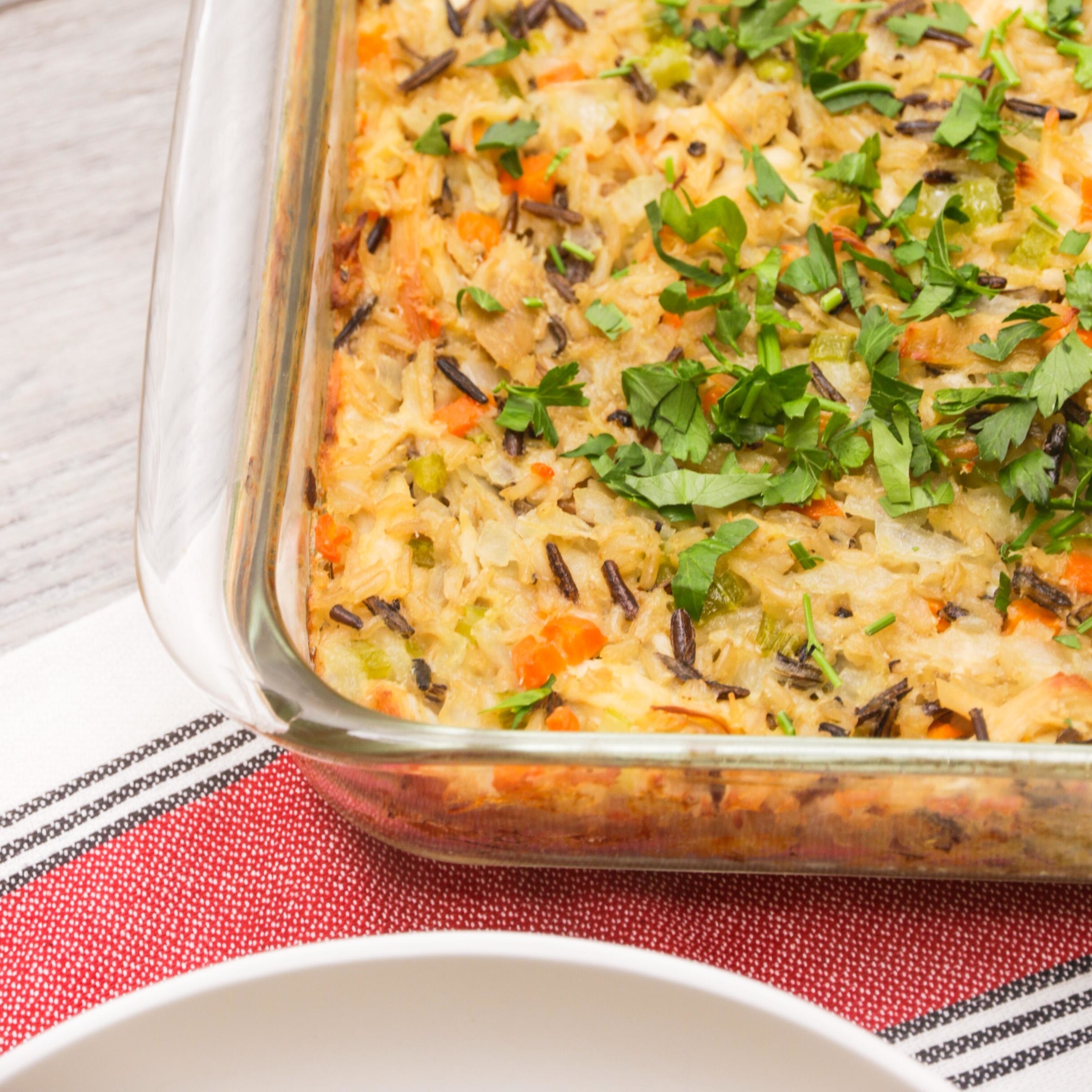 Close-up of a baked casserole dish with rice, vegetables, and parsley garnishing. A striped red and white towel and a white plate are placed beside the dish