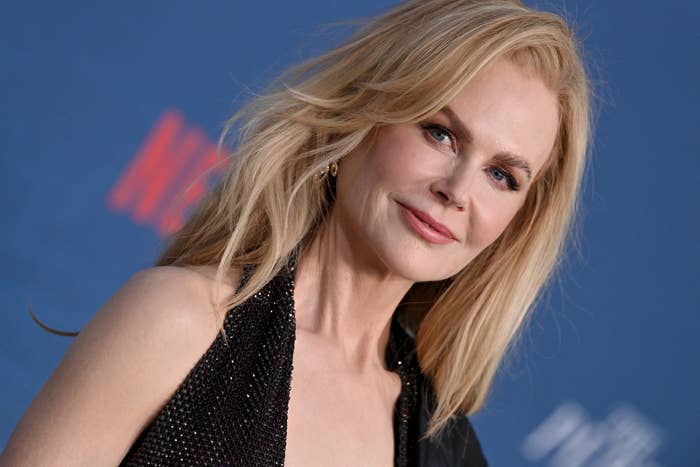 Nicole Kidman smiles, wearing a sleeveless black dress, with her blonde hair down, against a blurred blue background