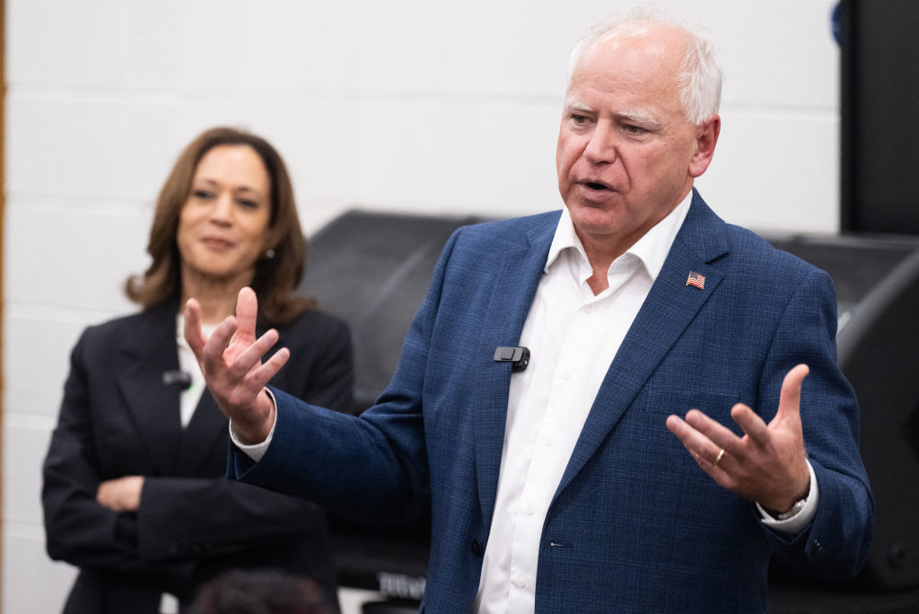 Kamala Harris stands in the background while Tim Walz speaks, gesturing with his hands, during a public event indoors