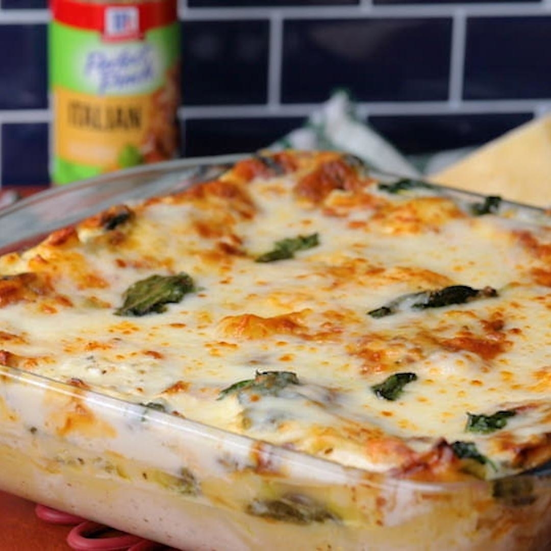 A freshly baked, cheesy lasagna in a glass baking dish is shown. It features layers of pasta, sauce, cheese, and spinach. A container of Italian seasoning is in the background