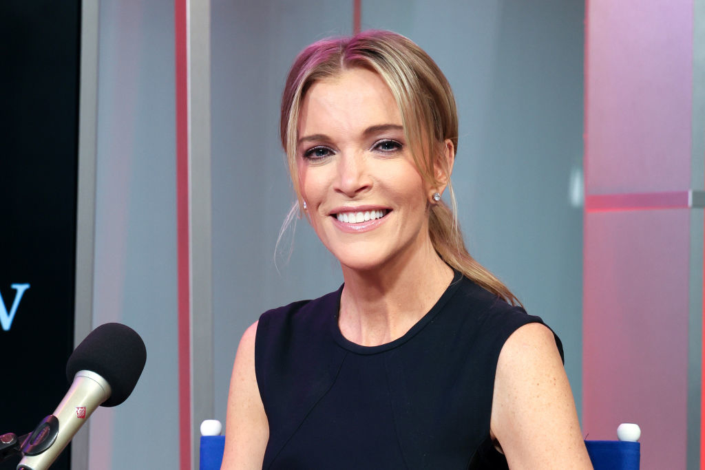 Megyn Kelly is sitting in a studio with a microphone in front of her, smiling and wearing a sleeveless, black dress