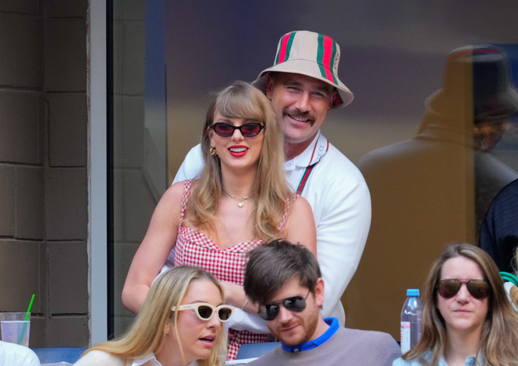 Taylor Swift wears a gingham dress while standing with Travis Kelce, who sports a striped bucket hat. They are behind two people seated, one in a beige top and sunglasses