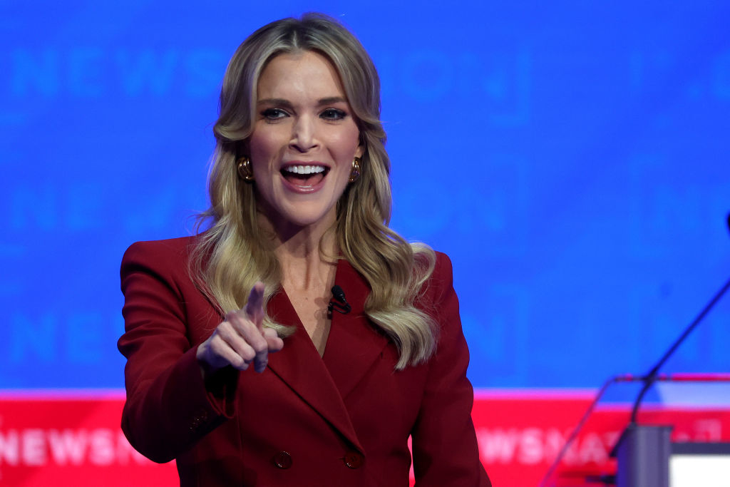 A smiling Megyn Kelly, wearing a formal blazer, gestures with her hand during an event. A podium with a microphone is visible in the background
