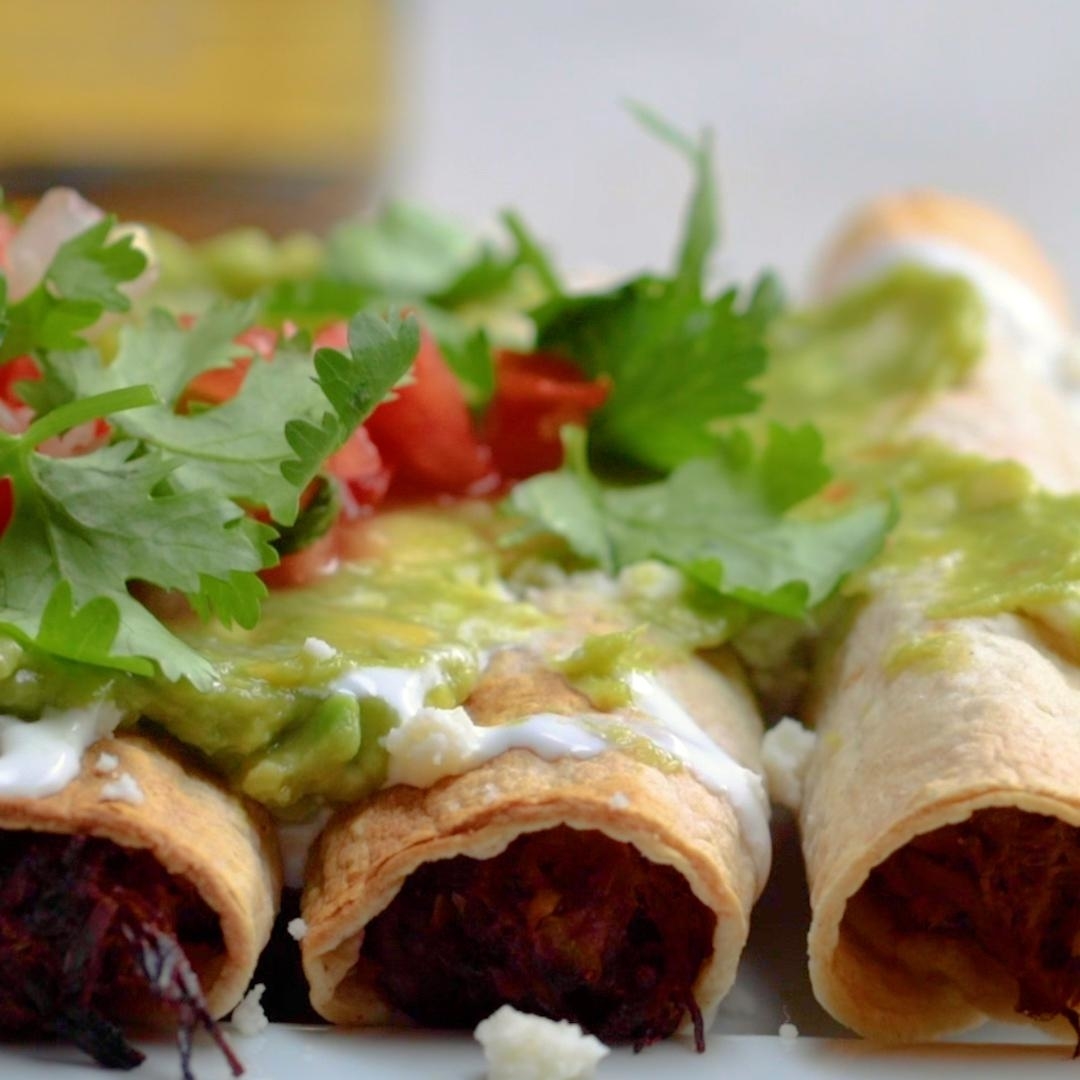 Close-up of three taquitos topped with fresh cilantro, diced tomatoes, and a creamy sauce