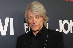 Jon Bon Jovi, in a black suit and shirt, stands in front of a backdrop with his name and other text partially visible. He smiles slightly at the camera