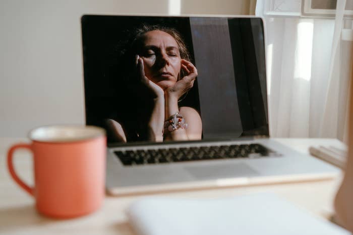 A laptop shows a woman with her eyes closed, resting her head on her hands. A pink cup is on the left side of the desk. The woman is not identified