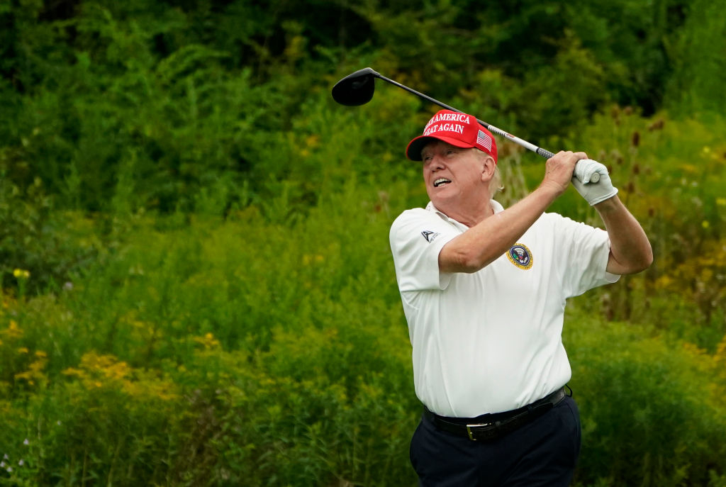 Donald Trump is swinging a golf club on a grassy golf course, wearing a red "Make America Great Again" hat and a white polo shirt