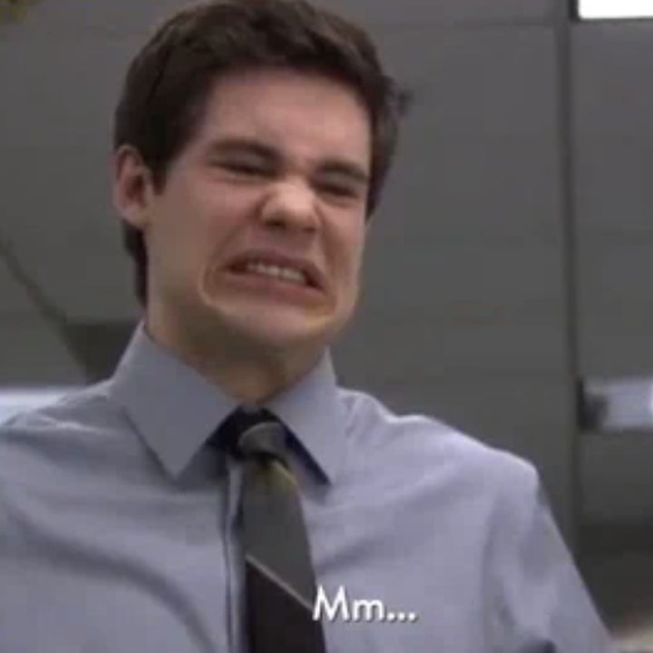 Adam DeVine grimaces while standing in an office, wearing a light gray shirt and dark tie. Subtitles at the bottom read, “Mm...”