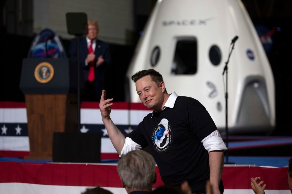 In the foreground, Elon Musk gestures while wearing a SpaceX shirt. In the background, Donald Trump stands at a podium with a space capsule displayed nearby