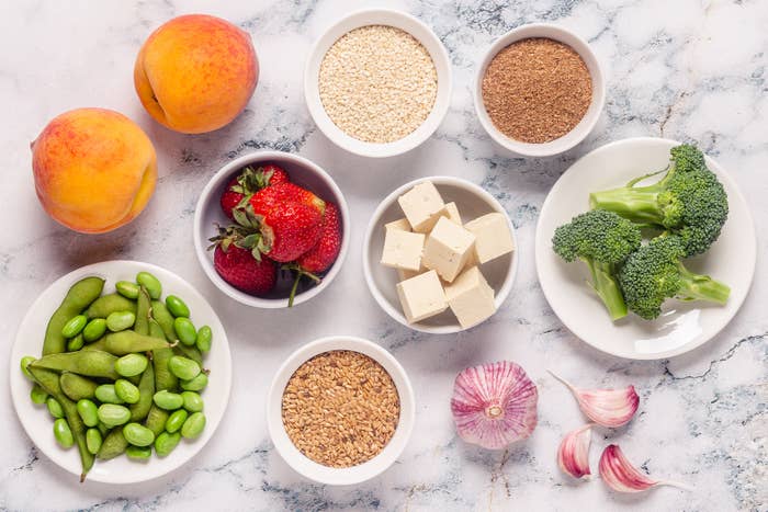 A variety of healthy foods are arranged on a marble surface, including peaches, strawberries, sesame seeds, wheat germ, broccoli, edamame, tofu, garlic, and flax seeds