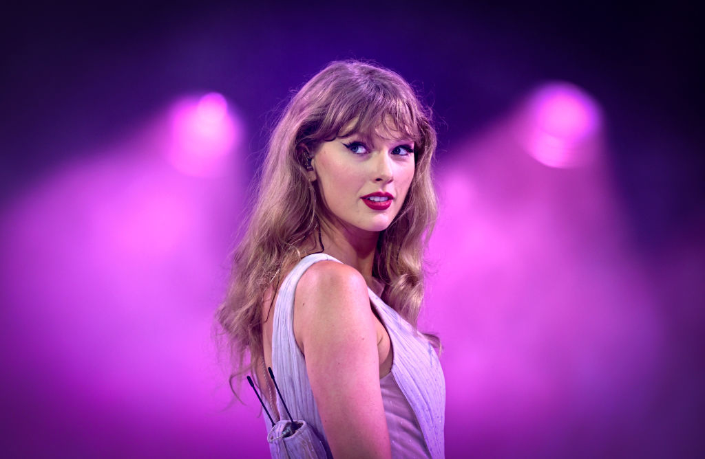 Taylor Swift is on stage in a sleeveless dress, with bright purple lights in the background, looking over her shoulder