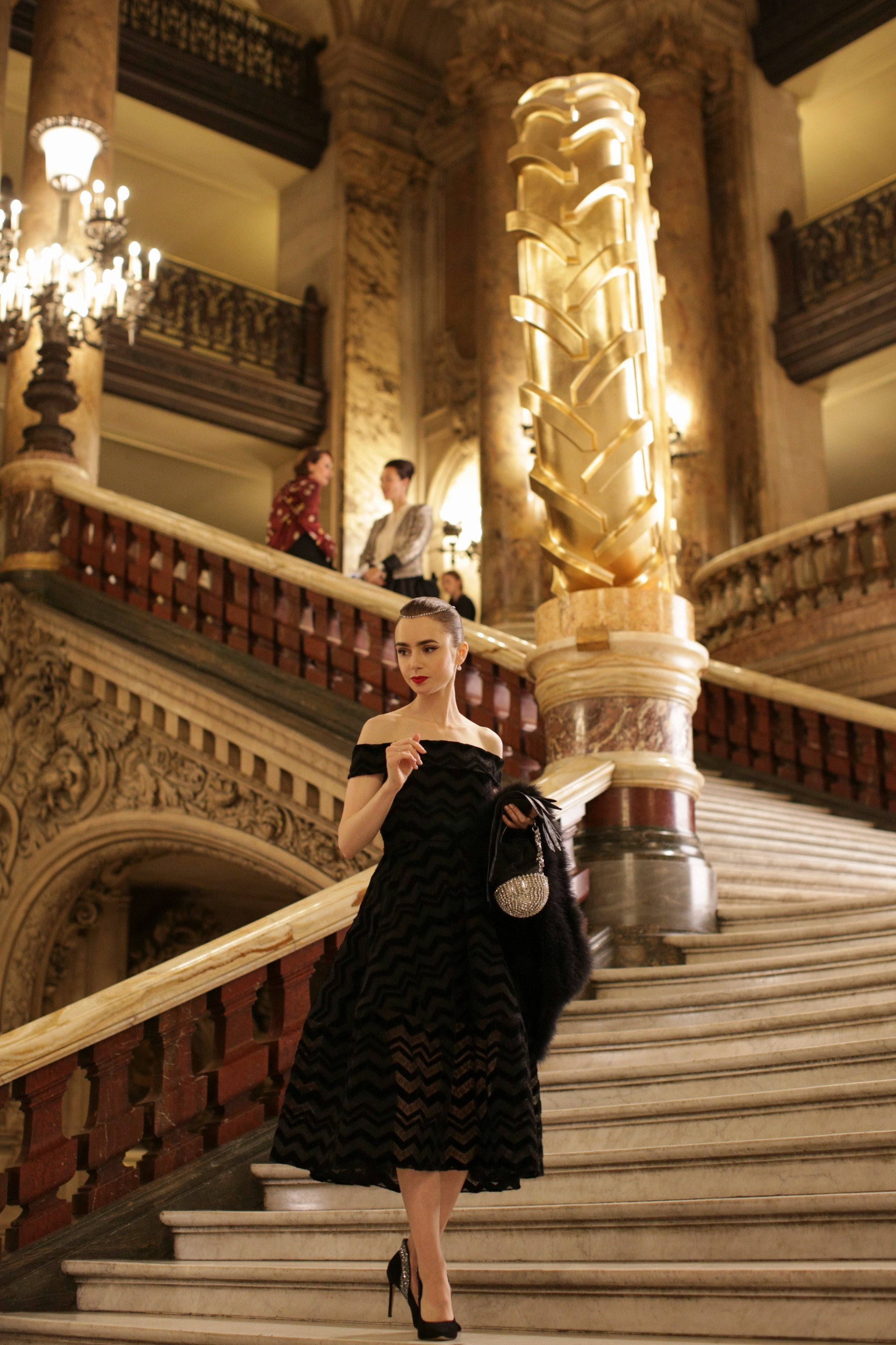 Lily Collins in a black off-the-shoulder dress ascends an elegant staircase with ornate decor. Another man and woman in stylish attire are in the background
