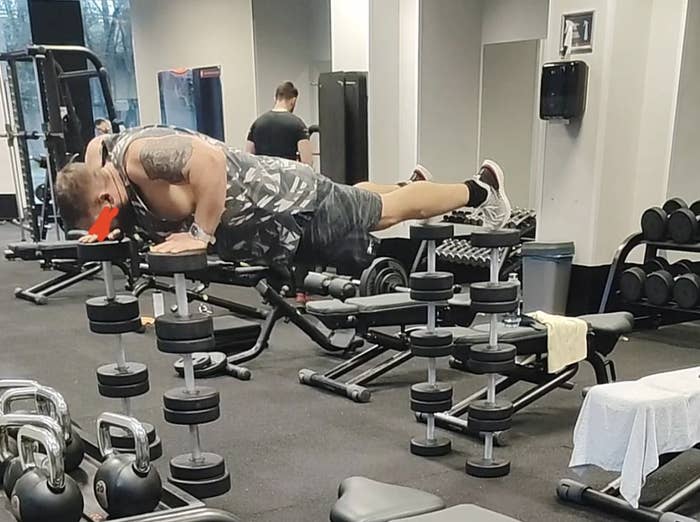 Man in a gym balancing on workout benches and weights, wearing a printed tank top and shorts, holding a red object in his mouth. Other gym-goers visible in background