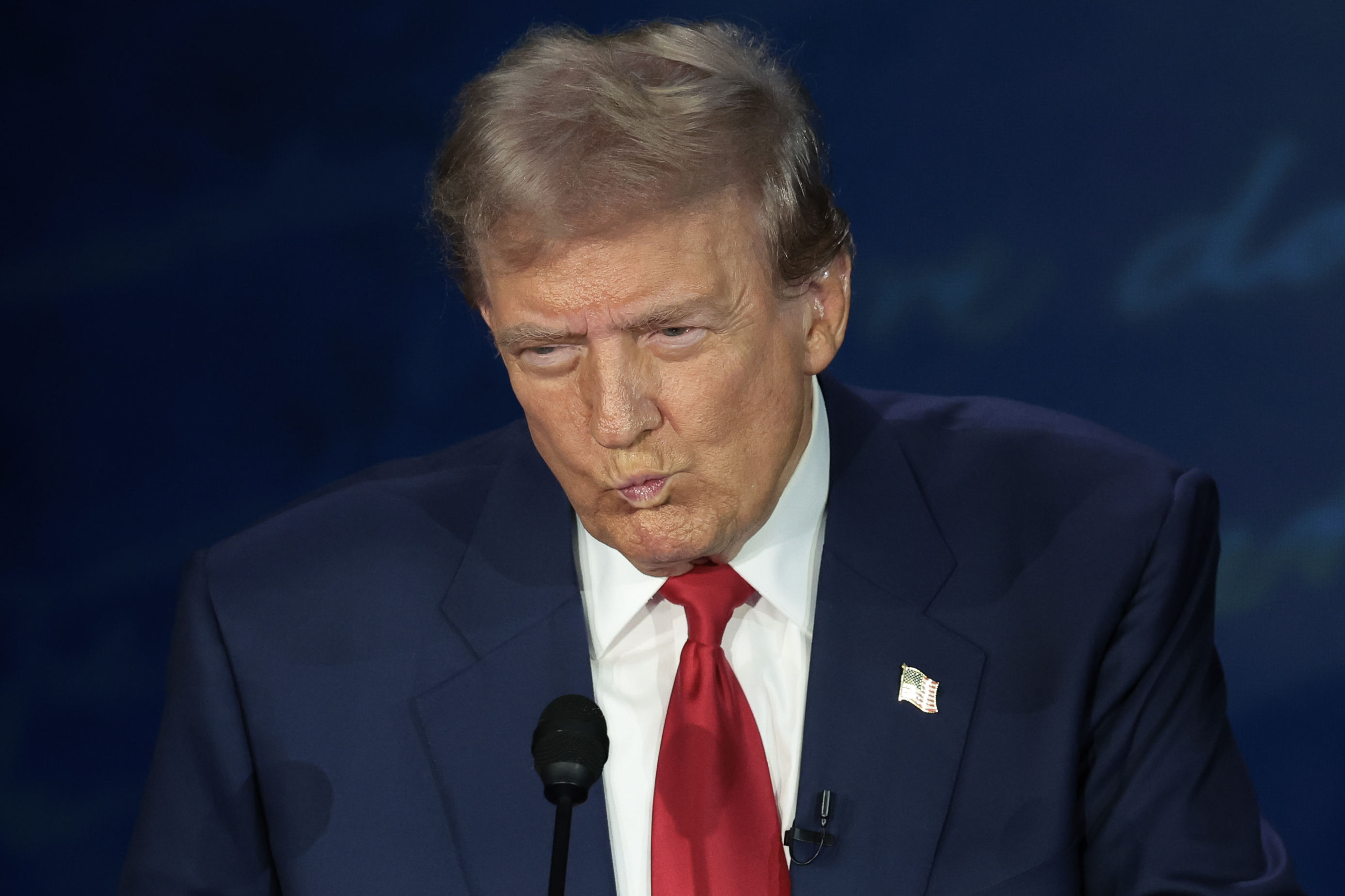 Donald Trump speaking at an event, wearing a suit with a white shirt and red tie, American flag pin on the lapel