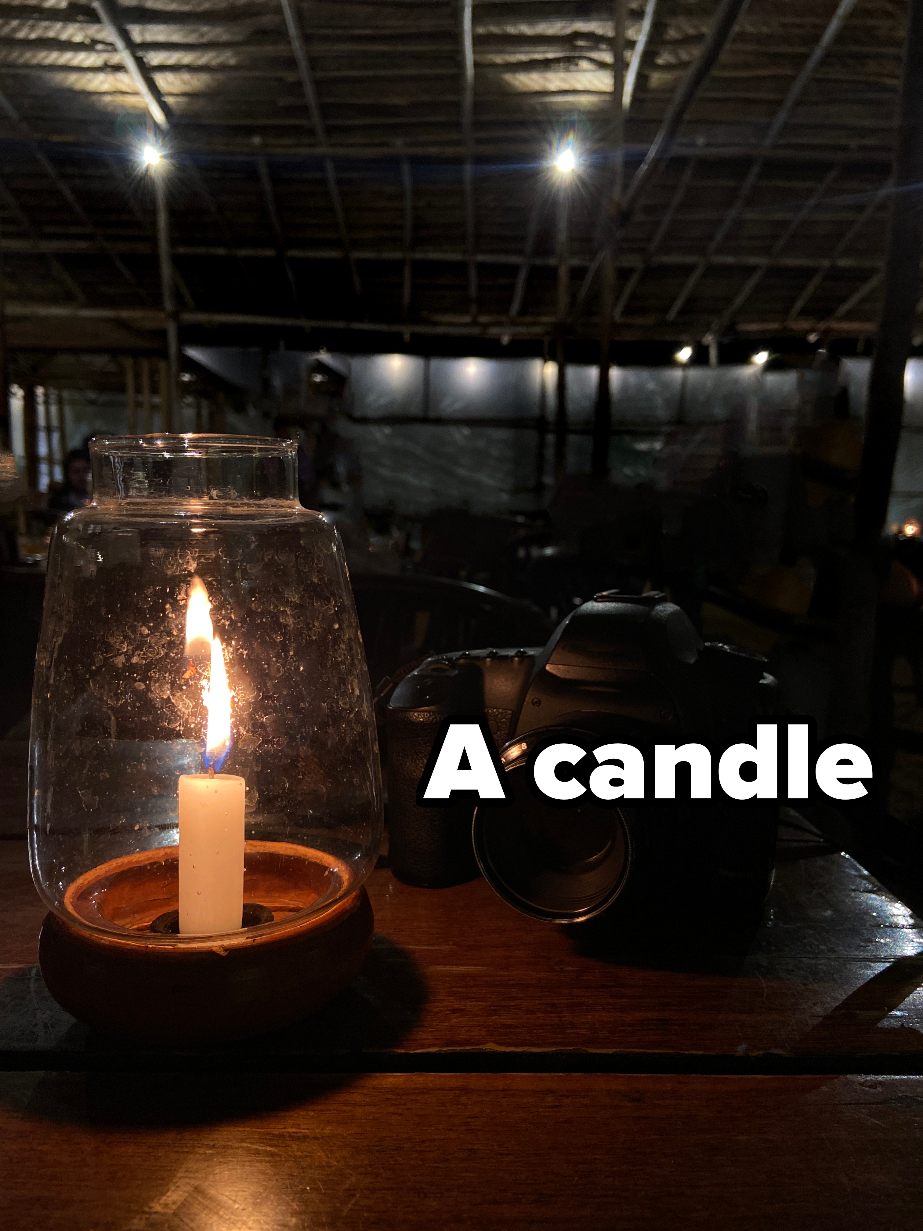 A lit candle in a glass holder and a camera on a wooden table in a dimly lit room