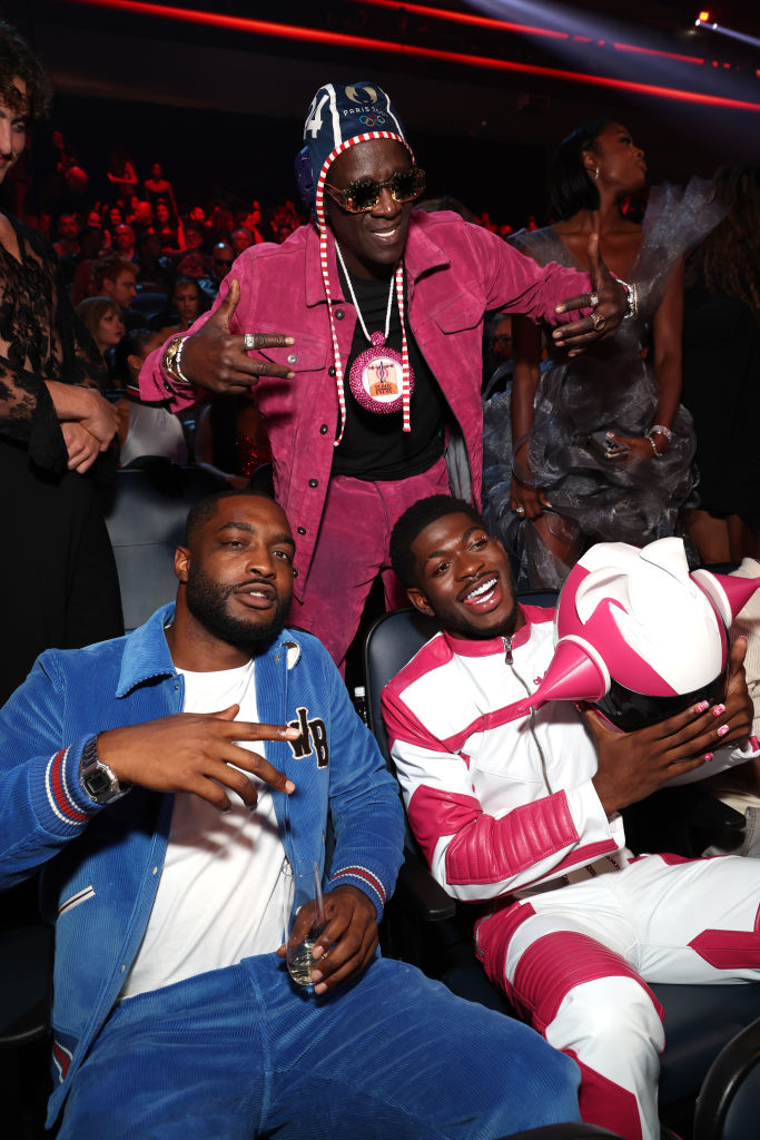 Flavor Flav, John Wall, and Lil Nas X pose together at an indoor event. Flavor Flav points and smiles behind Wall and Lil Nas X, who are seated and smiling