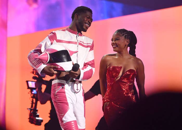 Lil Nas X and Halle Bailey smile at each other on stage. Lil Nas X wears a futuristic racing outfit holding a matching helmet. Halle Bailey wears a sparkling strapless dress