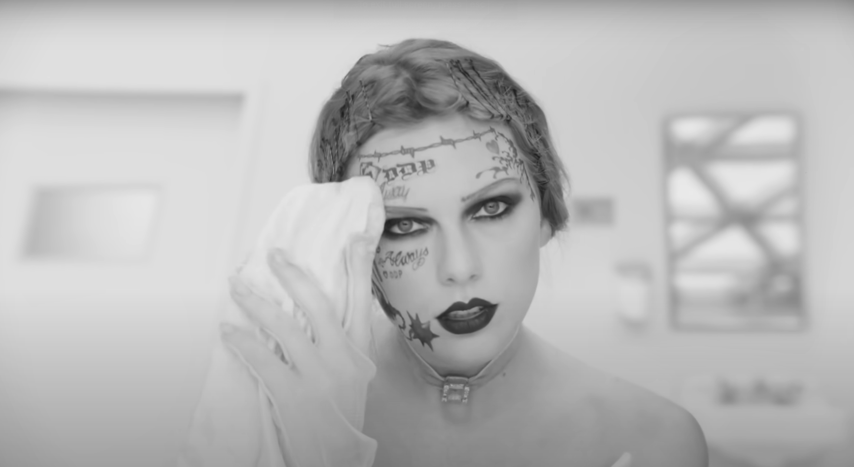 Taylor Swift in a stylized makeup look with tattoo-like drawings on her face, holding a cloth to her cheek