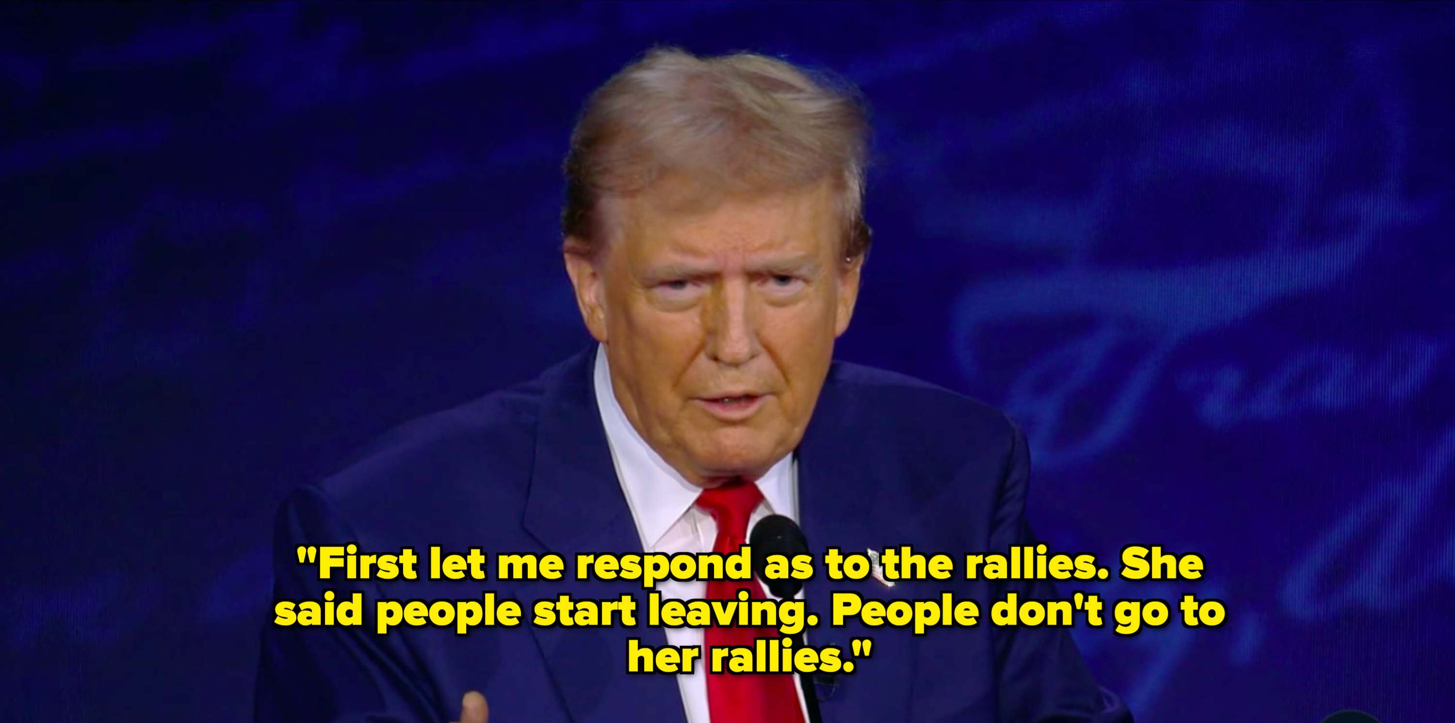 Donald Trump speaking at a podium with a microphone, wearing a suit and red tie