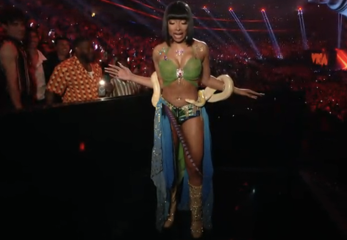 Megan Thee Stallion performing on stage wearing a green bikini top, blue skirt, and thigh-high boots, holding a large snake