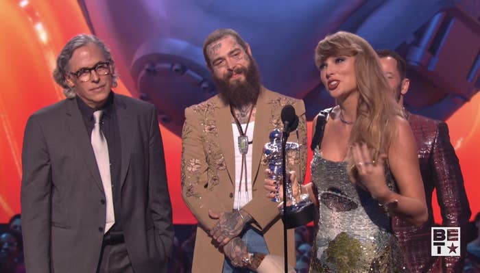 Taylor Swift holds a trophy and speaks at an award event, with Post Malone and others standing beside her