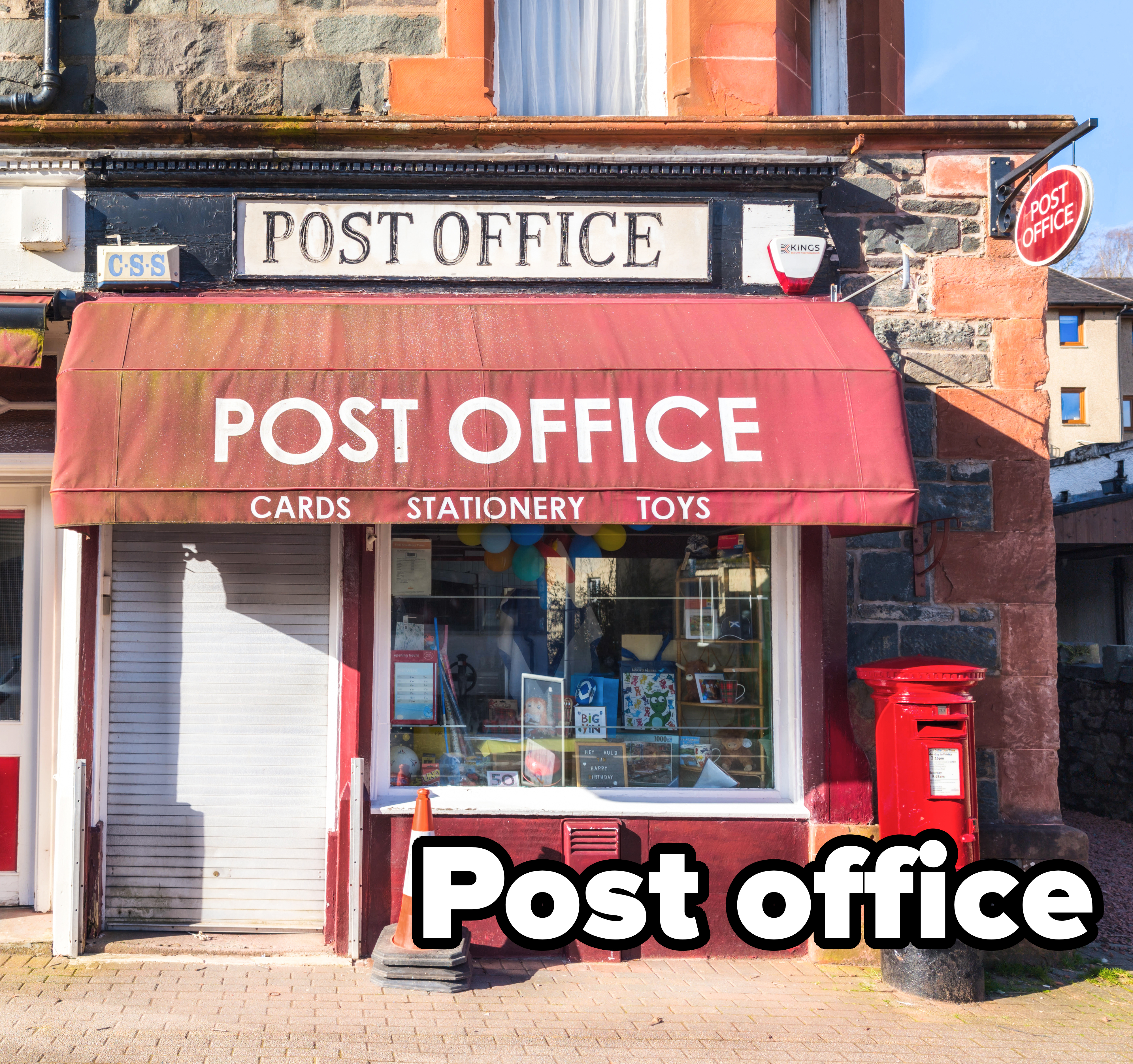 Post office storefront with signage for cards, stationery, and toys. A red mailbox and a construction cone are in front