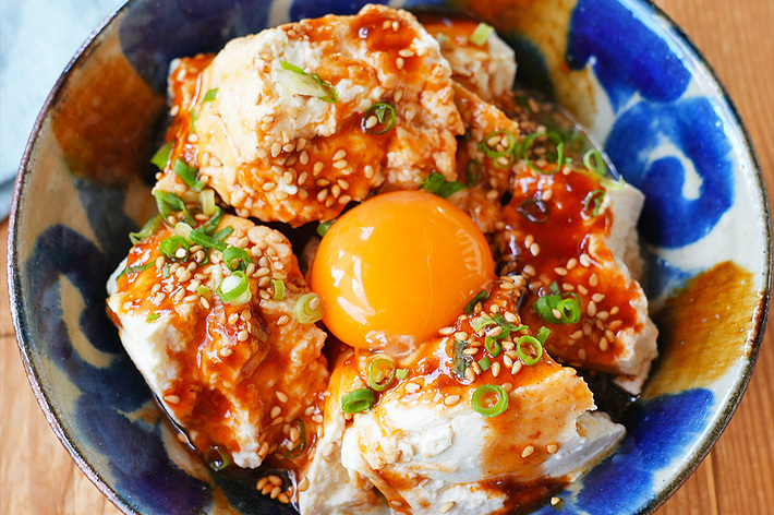 陶器の器に盛られた豆腐料理。中央には生卵の黄身が乗せられており、ネギやゴマが散らされています。