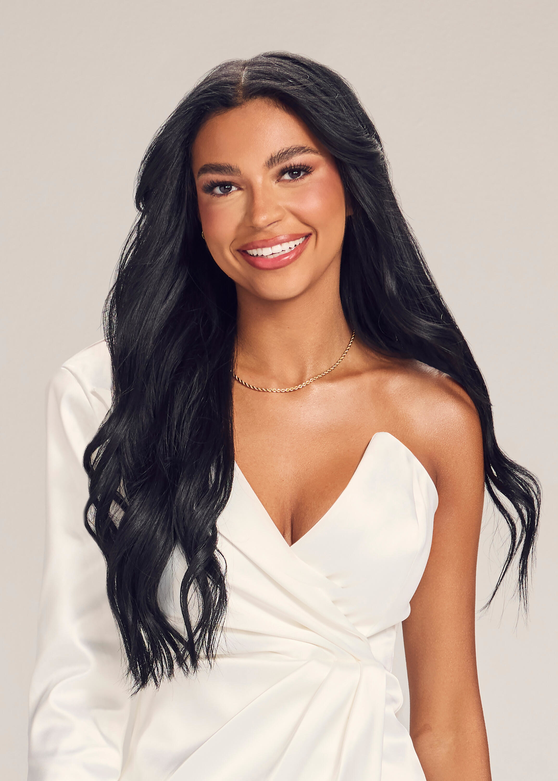 A woman in an elegant, off-shoulder white dress with long black hair and a gold necklace smiles for the camera