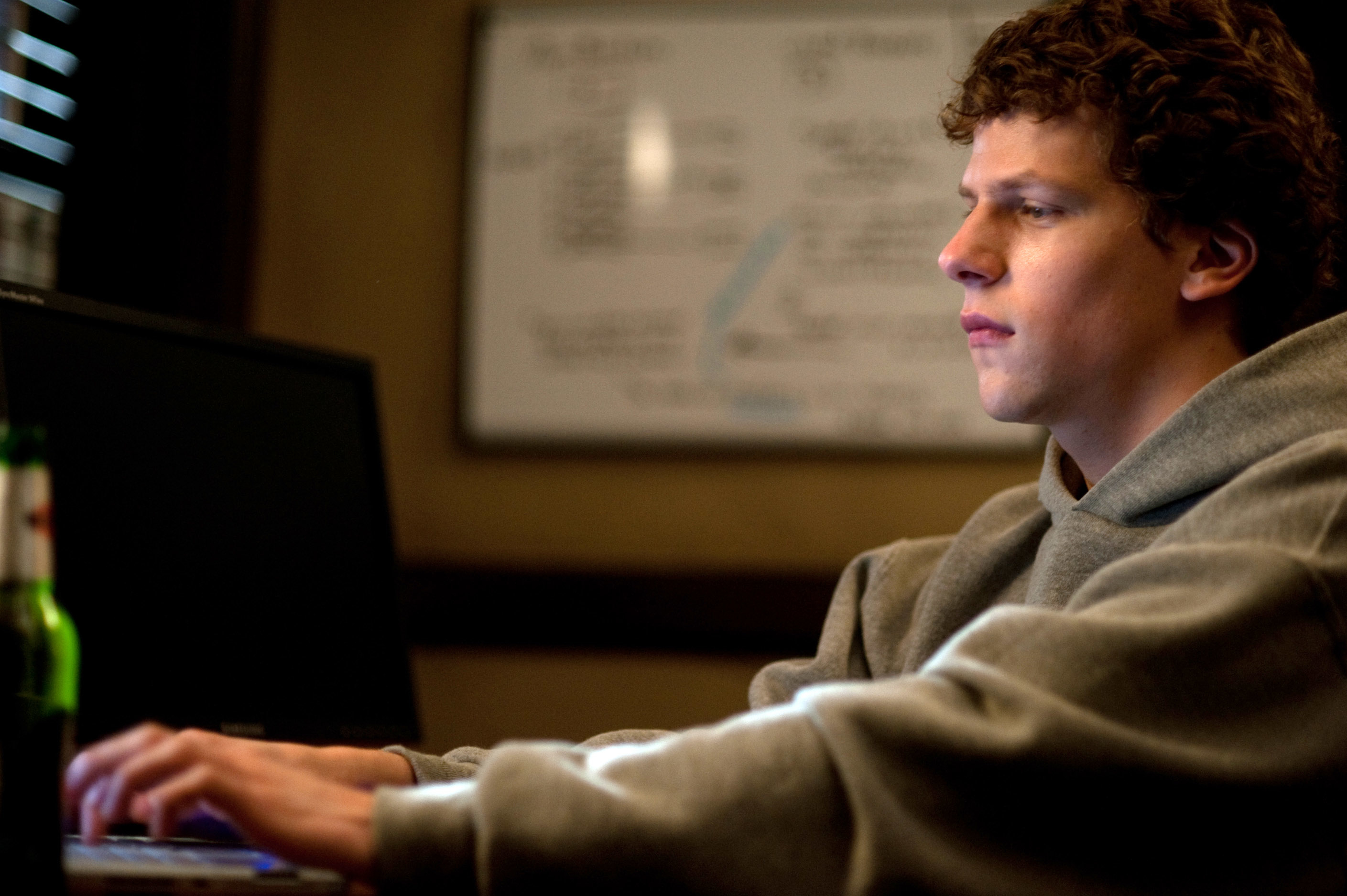 Jesse Eisenberg is sitting at a desk in a hoodie, focused on his laptop. A large whiteboard with writing is seen in the background