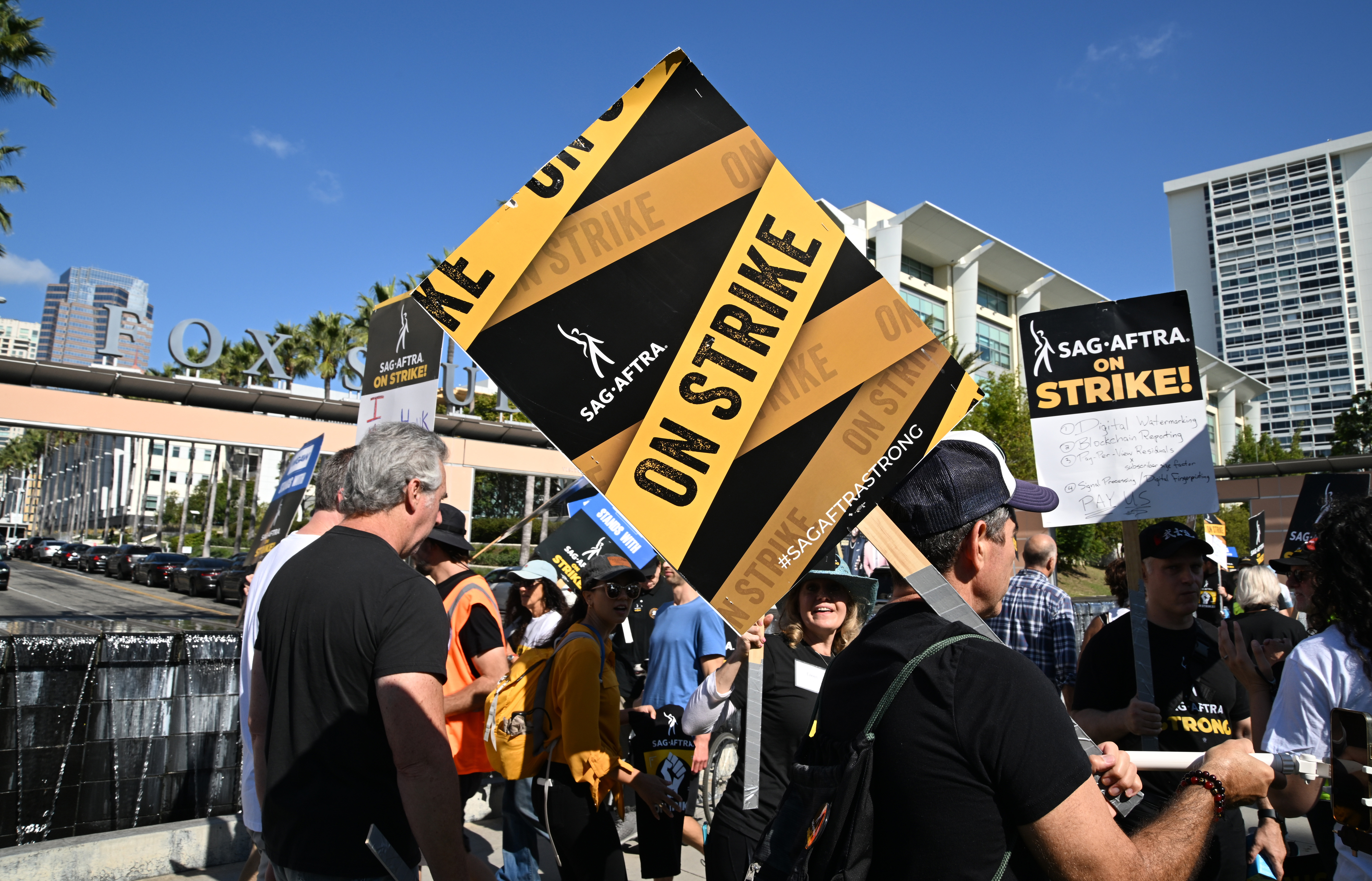 SAG-AFTRA members with protest signs demonstrate on the street in front of modern buildings, advocating for their rights during a strike