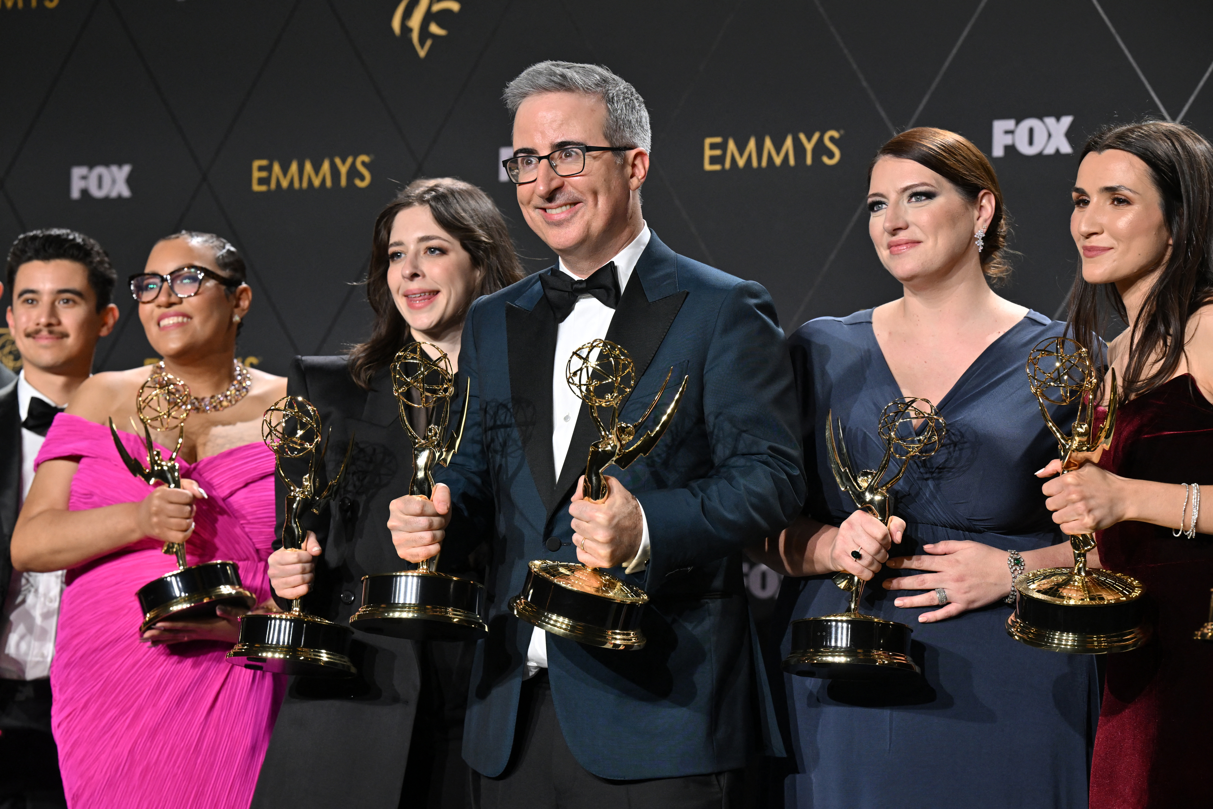 John Oliver, wearing a tuxedo, stands with other Emmy winners holding their awards